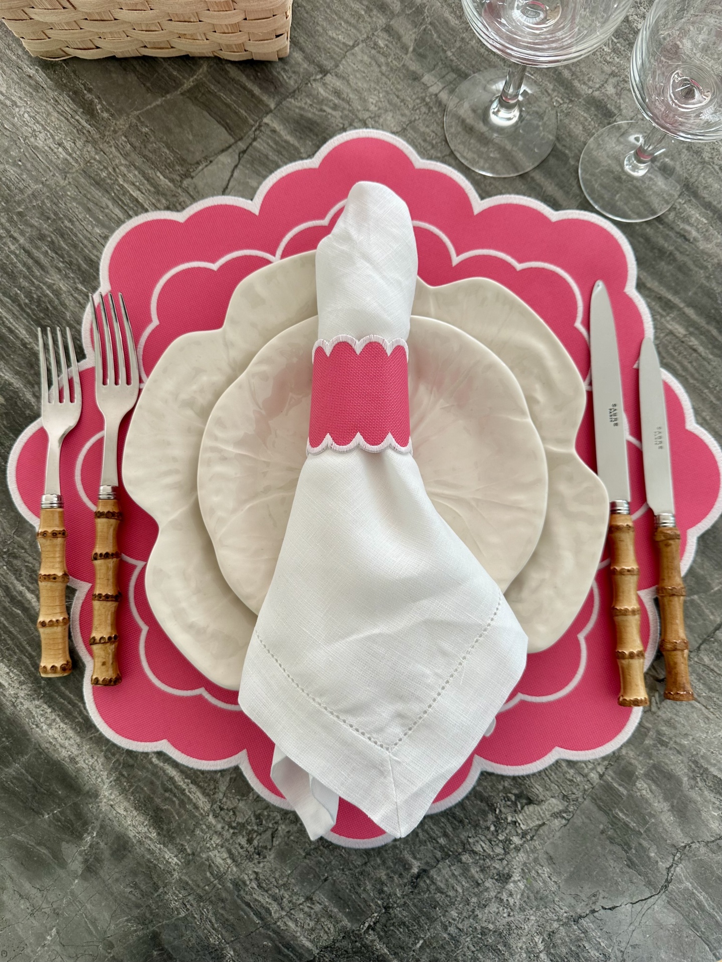 Mesa posta com prato branco, base rosa em forma de flor, guardanapo com argola, talheres com cabo de bambu e copos de vidro