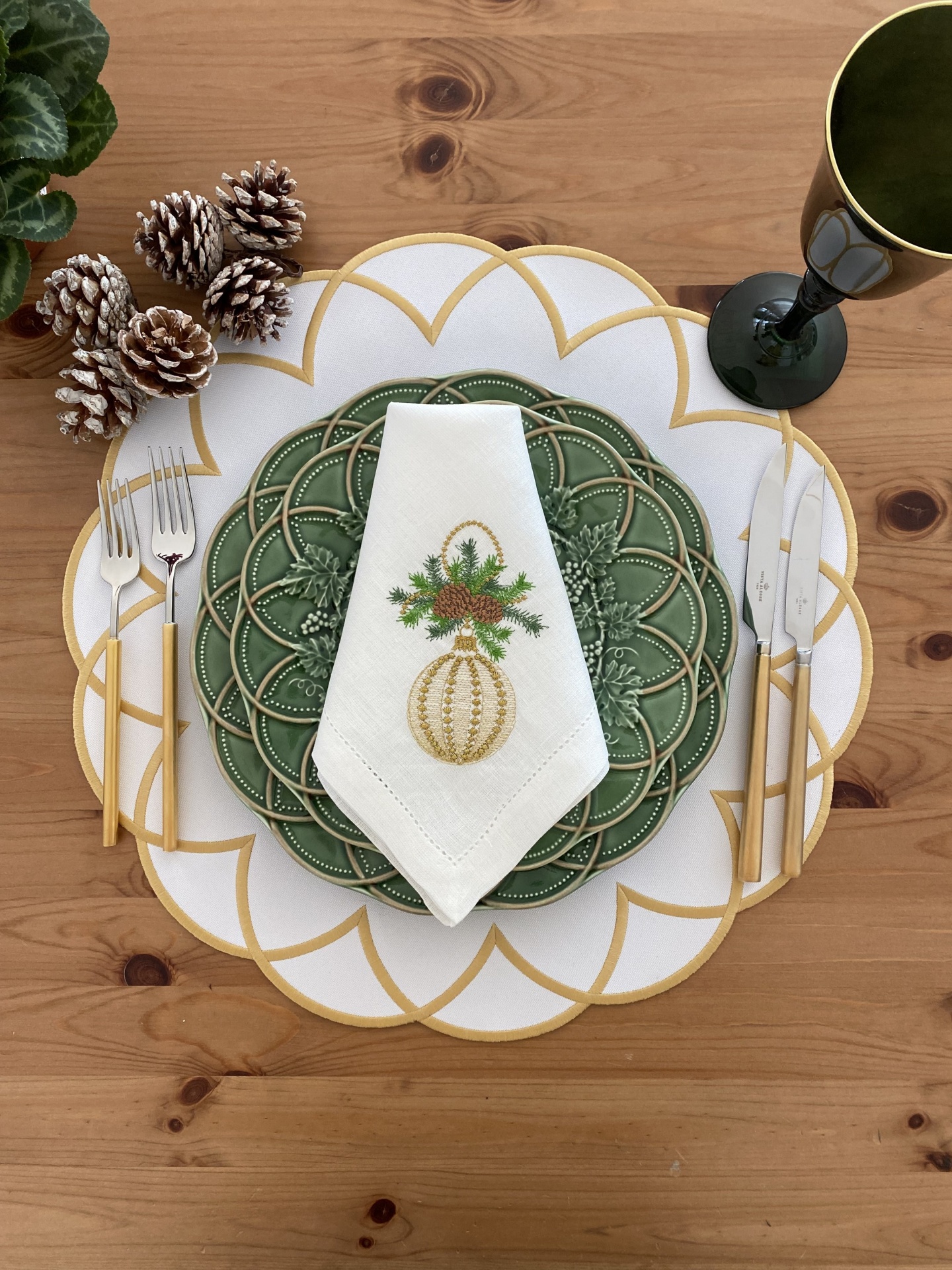 Mesa posta com prato verde, guardanapo branco bordado, talheres dourados, placemat branco e dourado, copo verde e pinhas decorativas.