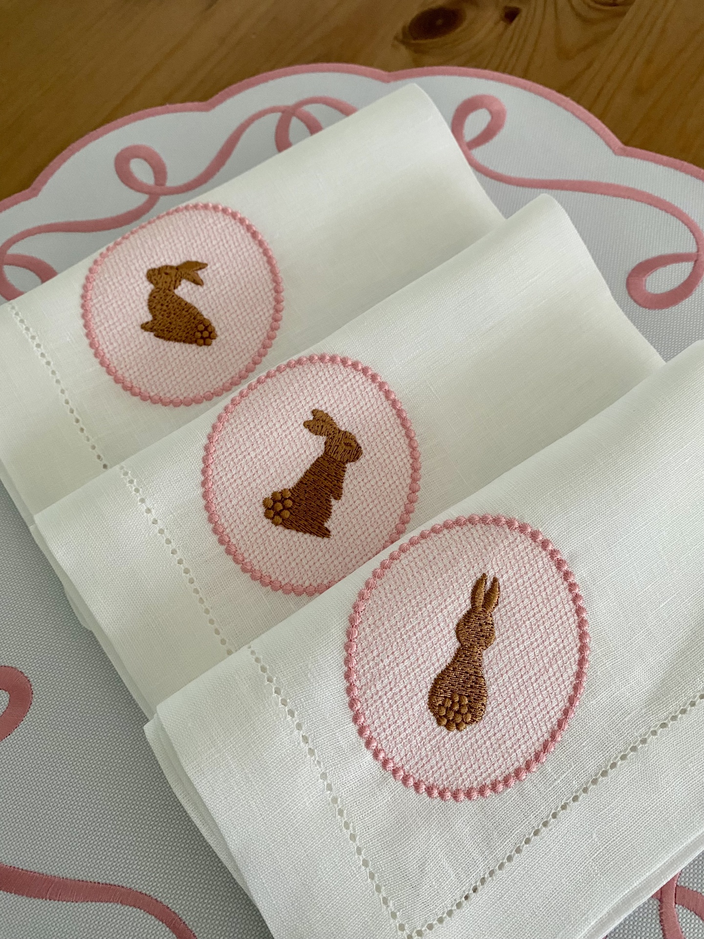 Guardanapos brancos com bordados de coelhos castanhos em oval cor de rosa sobre superfície branca com padrão rosa