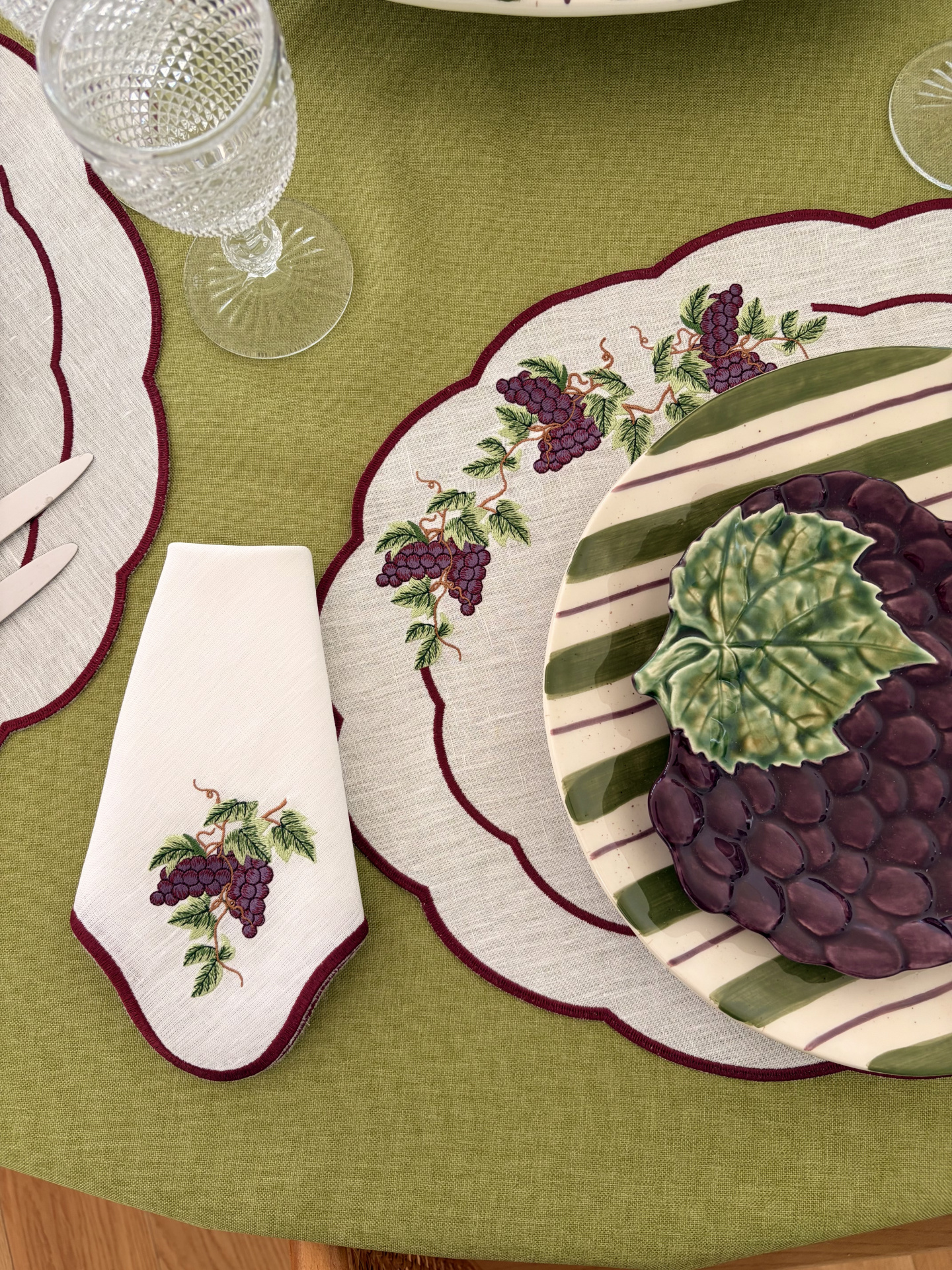 Conjunto de mesa com pratos decorados, guardanapo e jogo americano bordados, e copo de vidro texturizado sobre toalha verde.