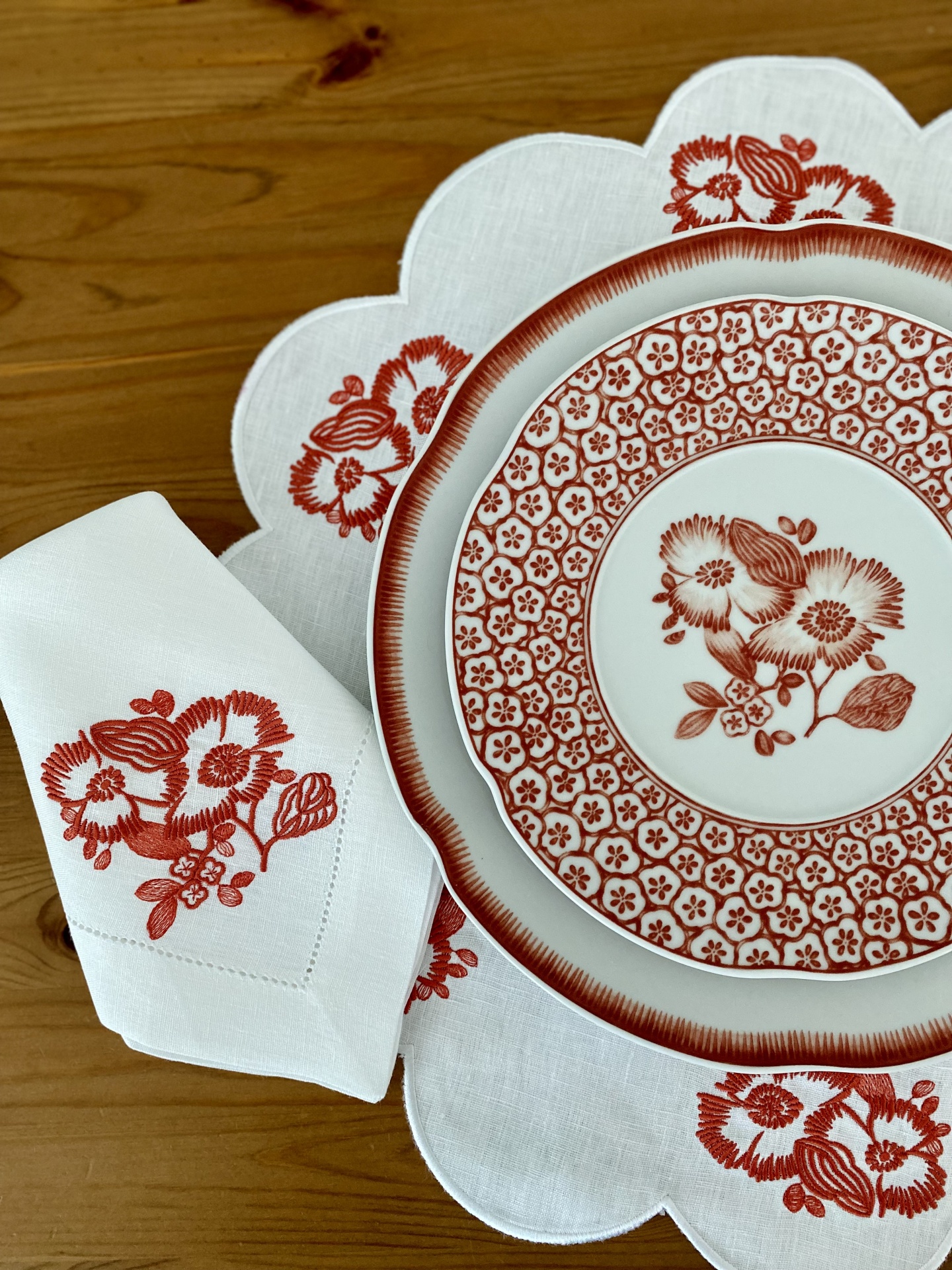 Conjunto de jantar com prato, guardanapo e toalha branca com padrões florais vermelhos sobre mesa de madeira