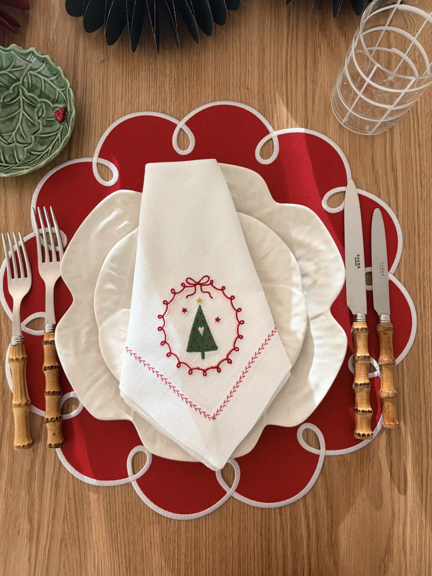 Conjunto de mesa com pratos brancos texturizados, guardanapo bordado de Natal, talheres com cabo em bambu, base de tecido vermelha e copo de vidro.