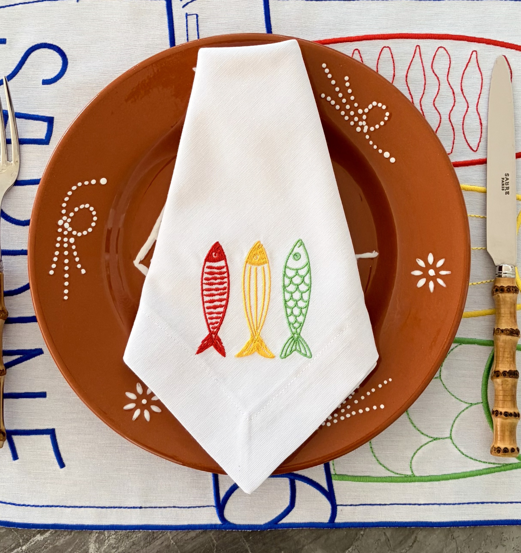 Conjunto de mesa com prato castanho decorado, guardanapo branco bordado com peixes multicoloridos, faca com cabo de madeira e toalha bordada colorida