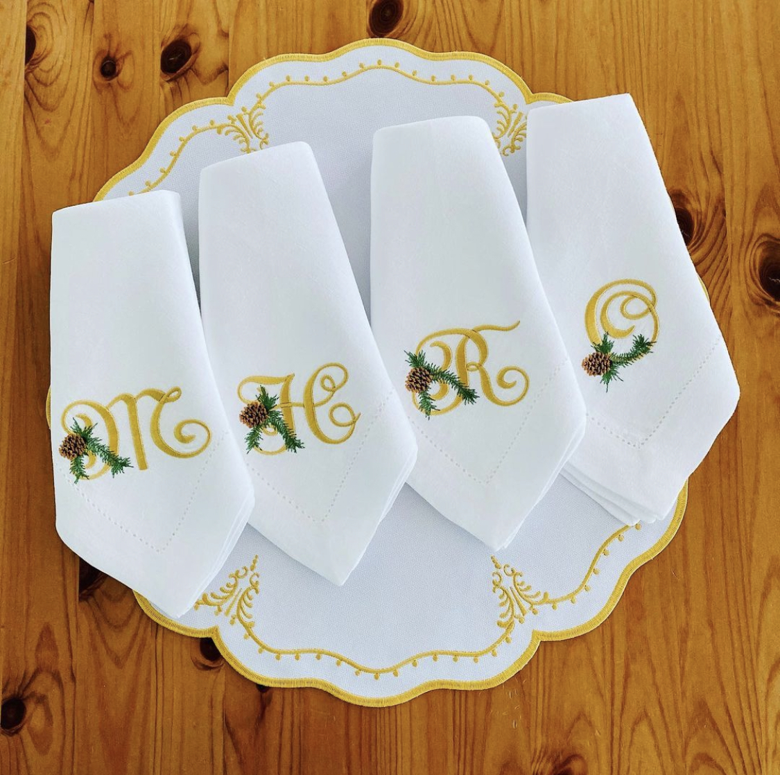 Guardanapos brancos bordados com letras douradas e decoração verde e castanha, sobre prato branco com detalhes dourados e madeira.