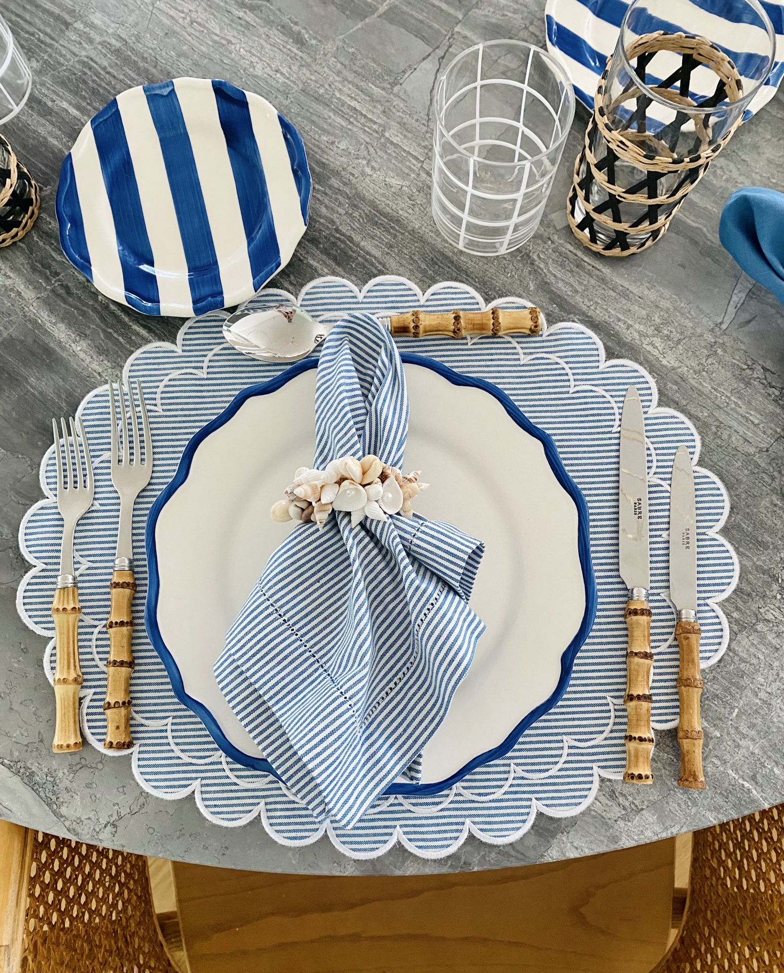 Mesa posta com prato branco e azul, prato pequeno às riscas, talheres com cabo de bambu, guardanapo azul e base de prato decorativa