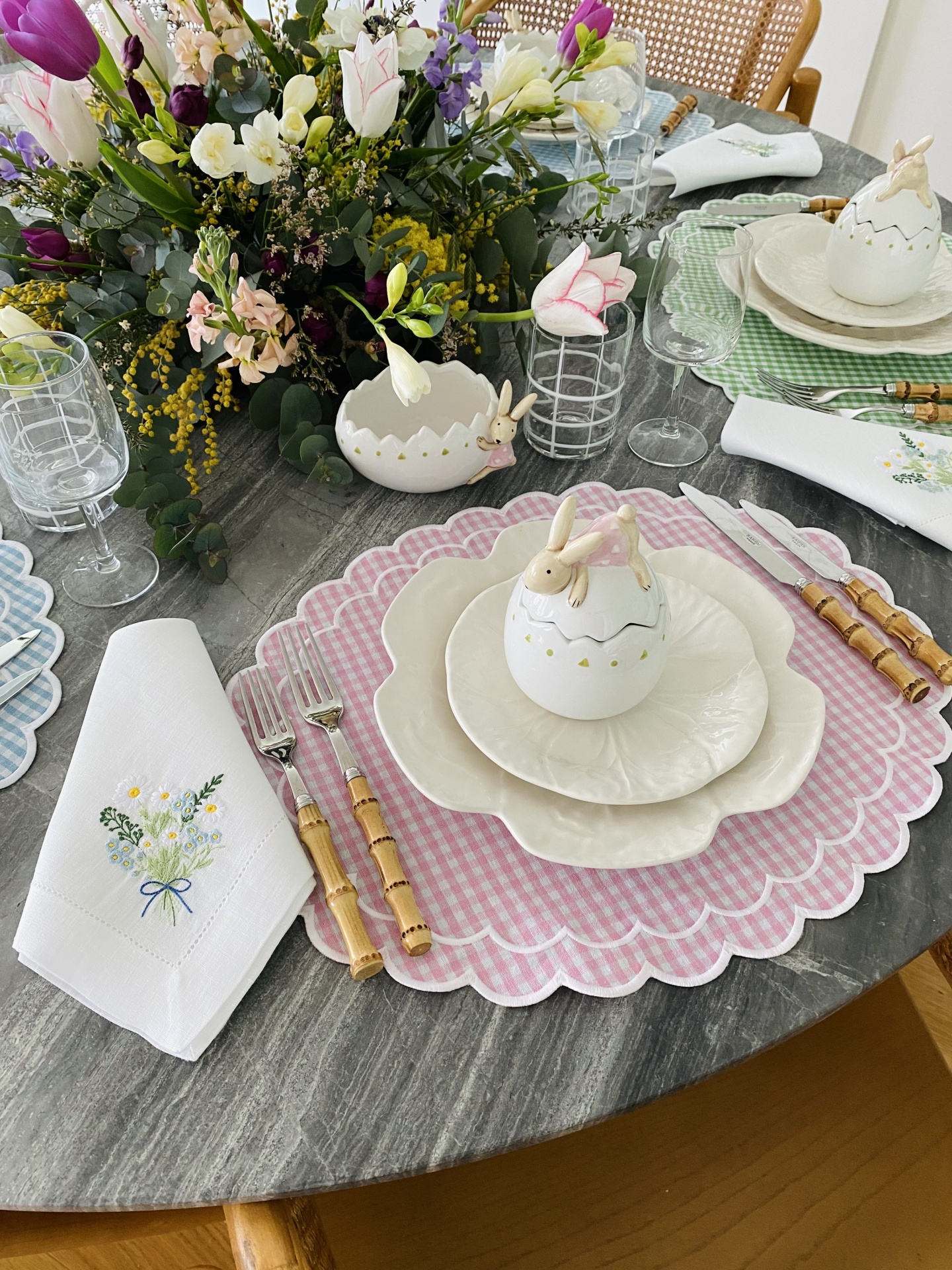 Mesa com jogo americano rosa xadrez, prato em forma de flor, ovo decorativo com coelho, talheres de cabo madeira e jarro de flores.