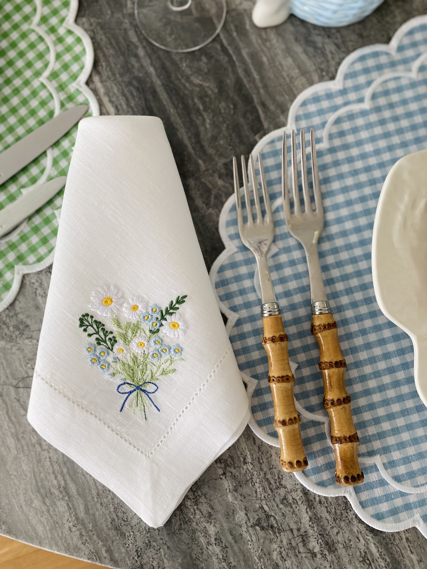 Guardanapo branco bordado com flores ao lado de garfos com cabo em bambu e prato sobre sousplat azul xadrez