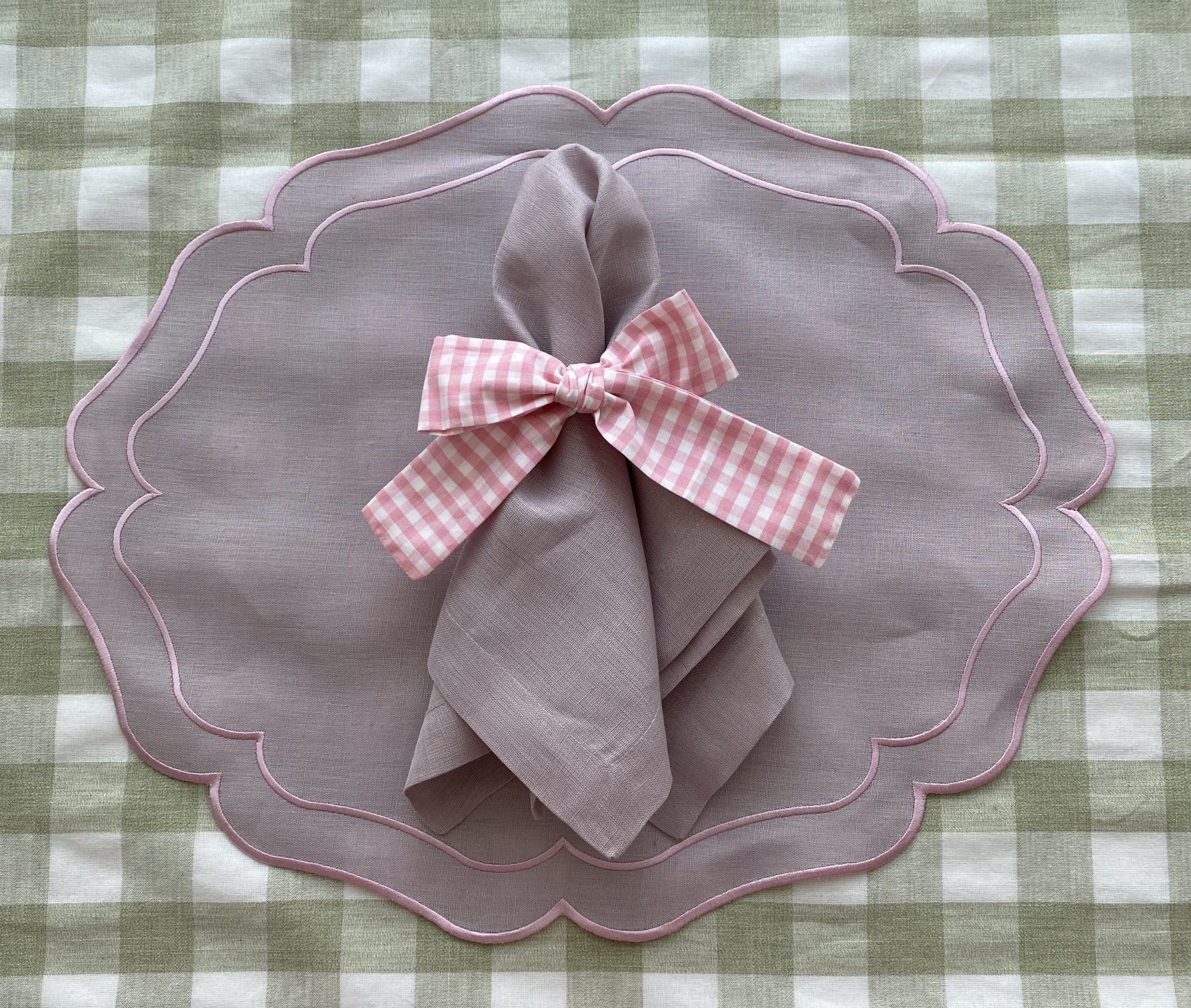 Guardanapo cinza com laço rosa às riscas sobre individual rosa ondulado numa toalha xadrez verde e branco.