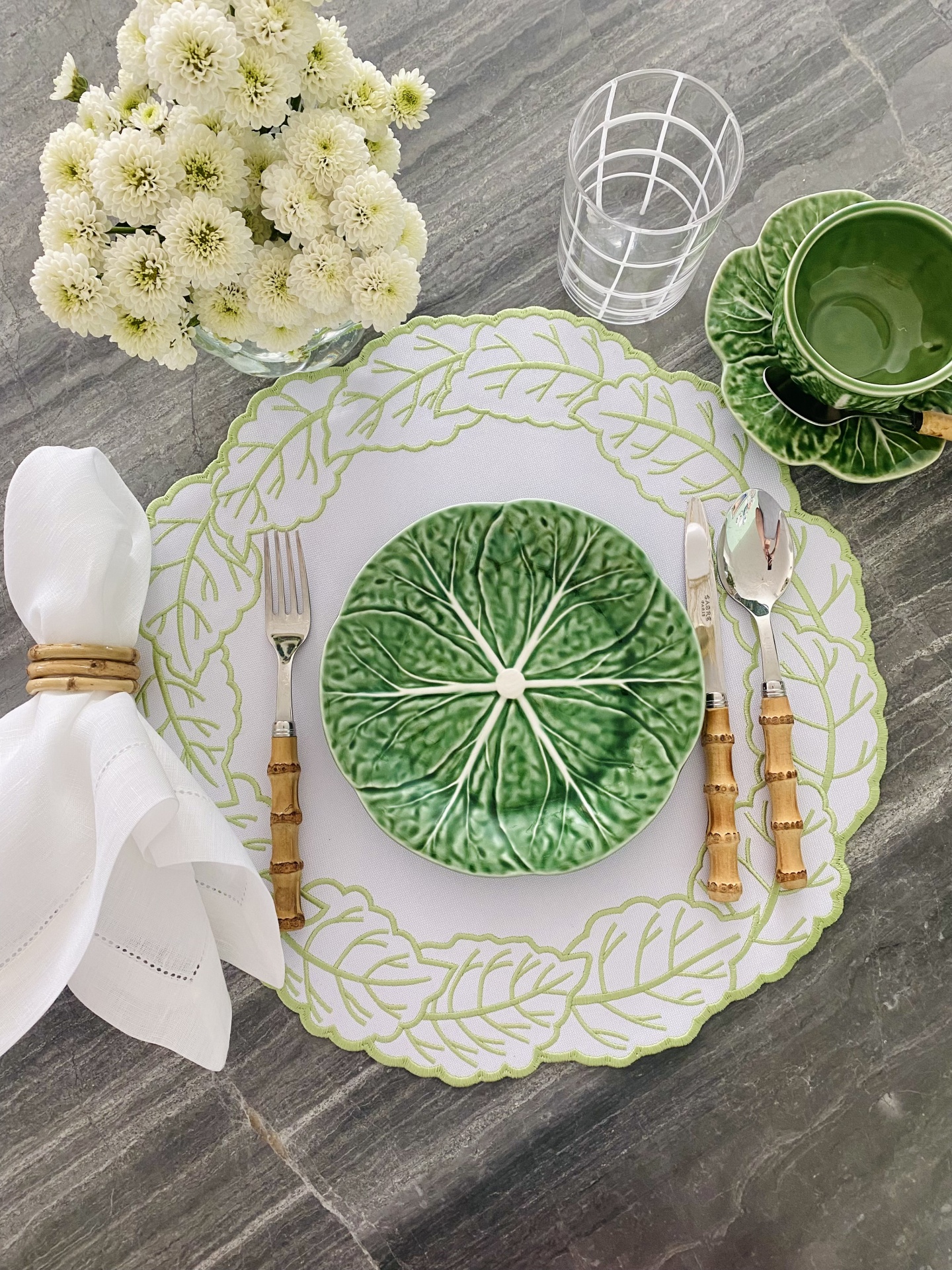 Mesa posta com prato verde de folha, prato branco com folhas desenhadas, talheres com cabo de bambu, guardanapo branco e vaso com flores brancas