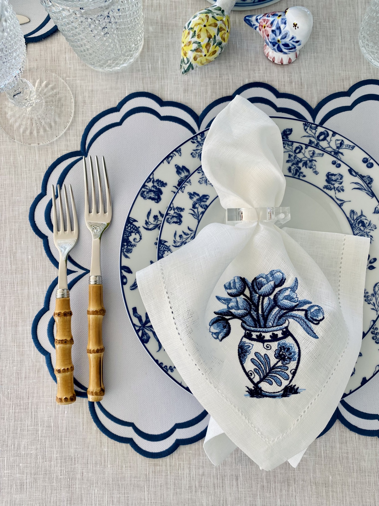 Mesa posta com prato branco e azul, talheres com cabos de bambu, guardanapo branco bordado e copos de vidro