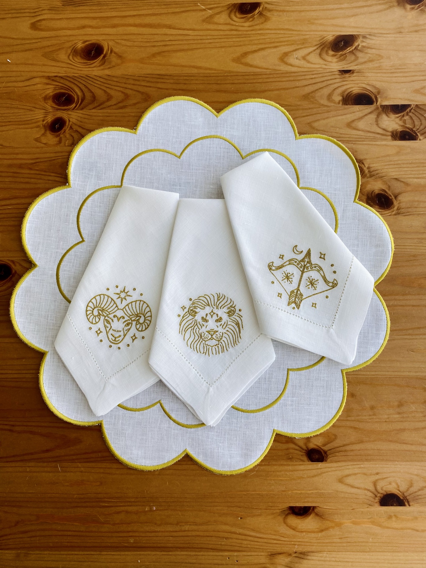 Três guardanapos brancos bordados com signos do zodíaco sobre passadeira branca com borda amarela numa mesa de madeira.
