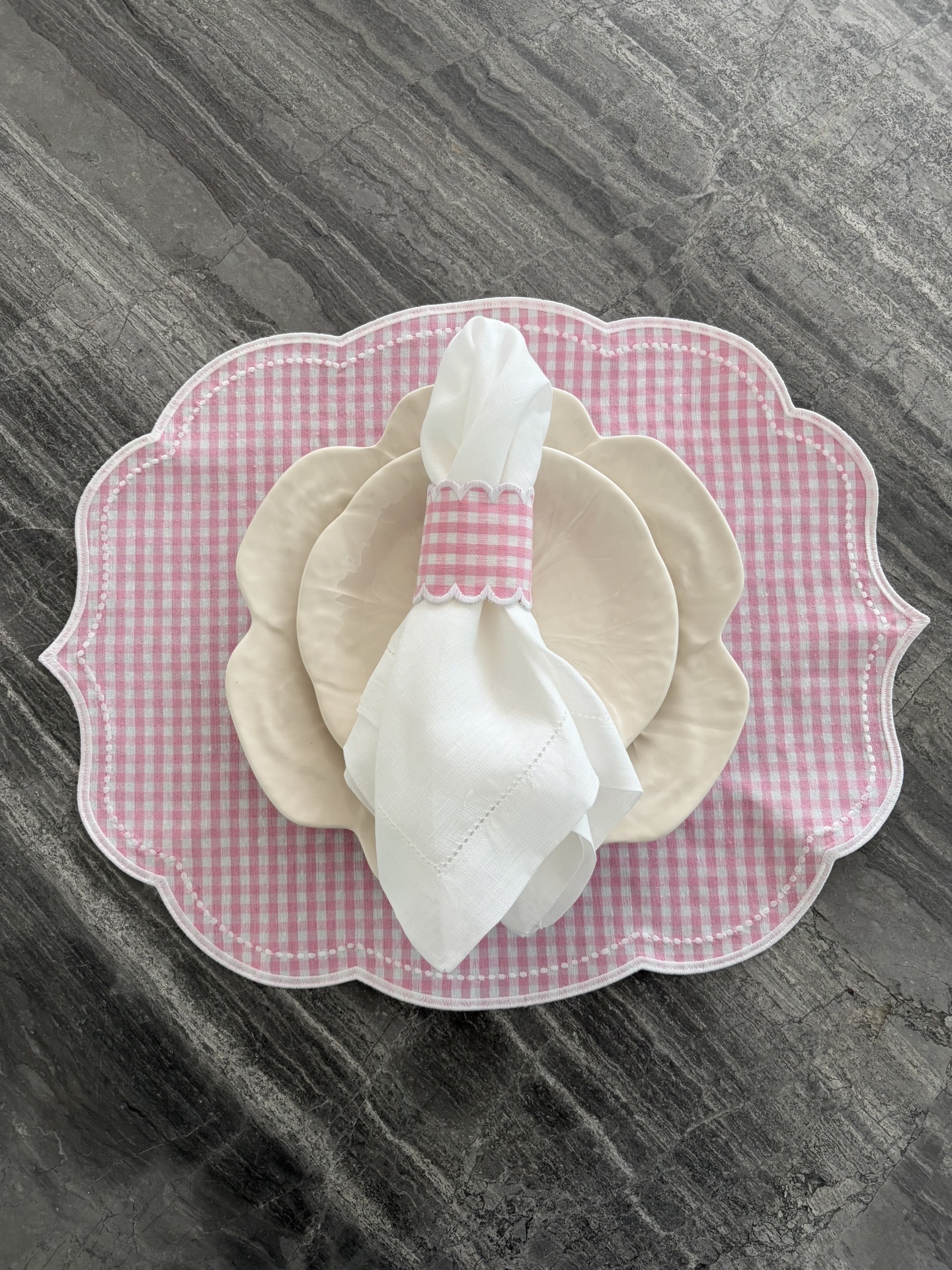 Jogo de mesa com pratos cerâmicos creme, guardanapo branco com argola xadrez rosa e branco, individual têxtil rosa xadrez em superfície cinzenta