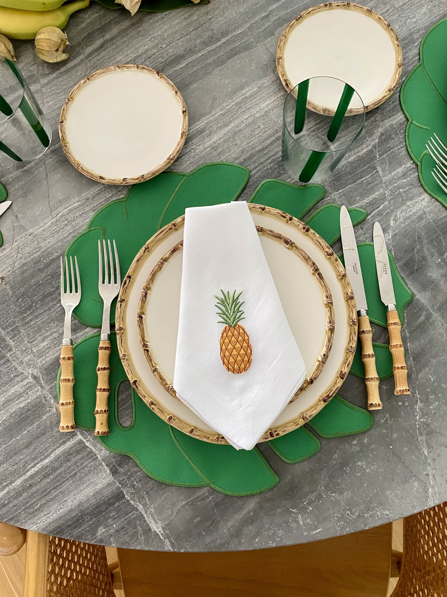 Conjunto de louça e talheres com padrão de bambu e acessório decorativo de ananás numa mesa cinza.