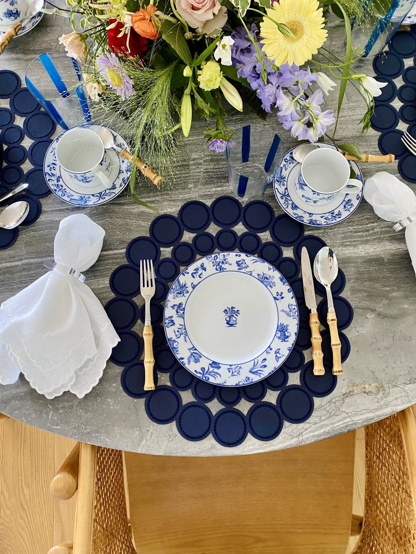 Mesa decorada para refeição com pratos brancos e azul, talheres com cabos de bambu, guardanapos brancos e arranjo floral colorido.