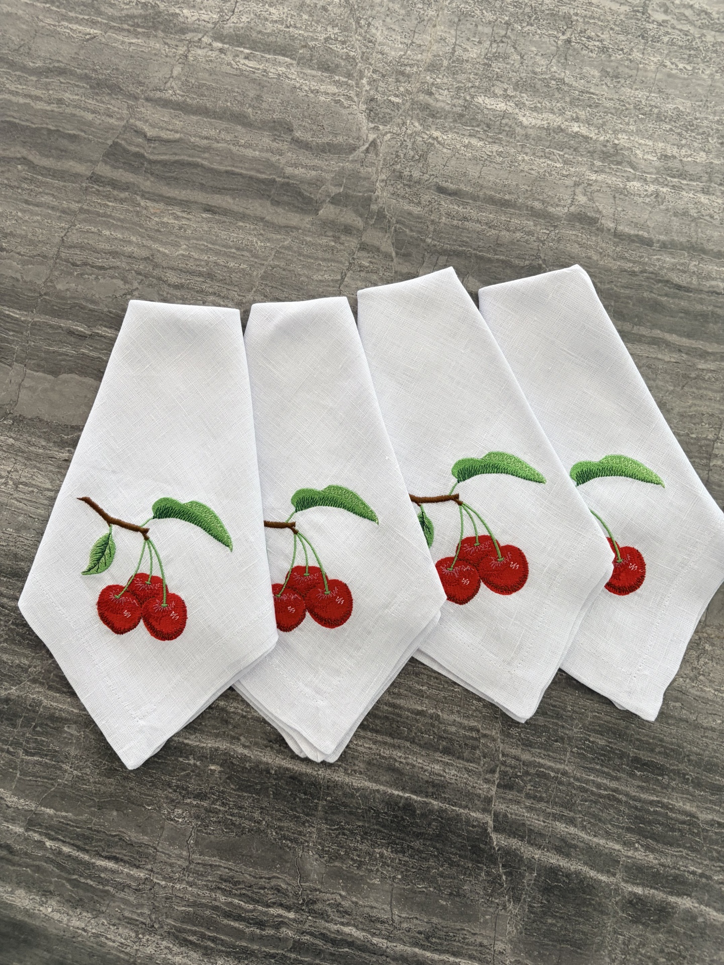 Guardanapos brancos bordados com cerejas vermelhas e folhas verdes sobre superfície cinza.