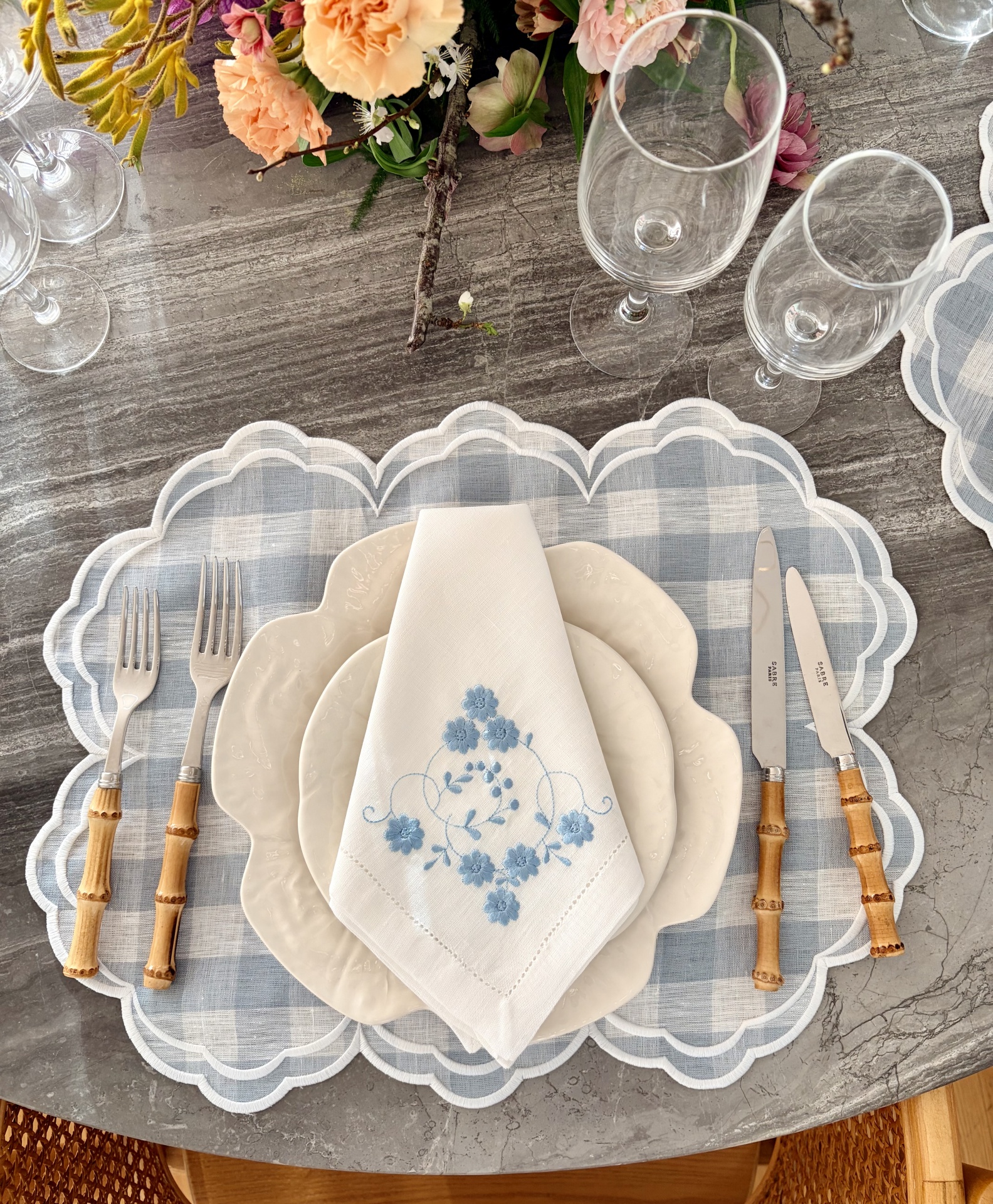 Conjunto de jantar com pratos brancos, guardanapo bordado azul, talheres com cabo de bambu, copos de vidro e base de tecido azul xadrez