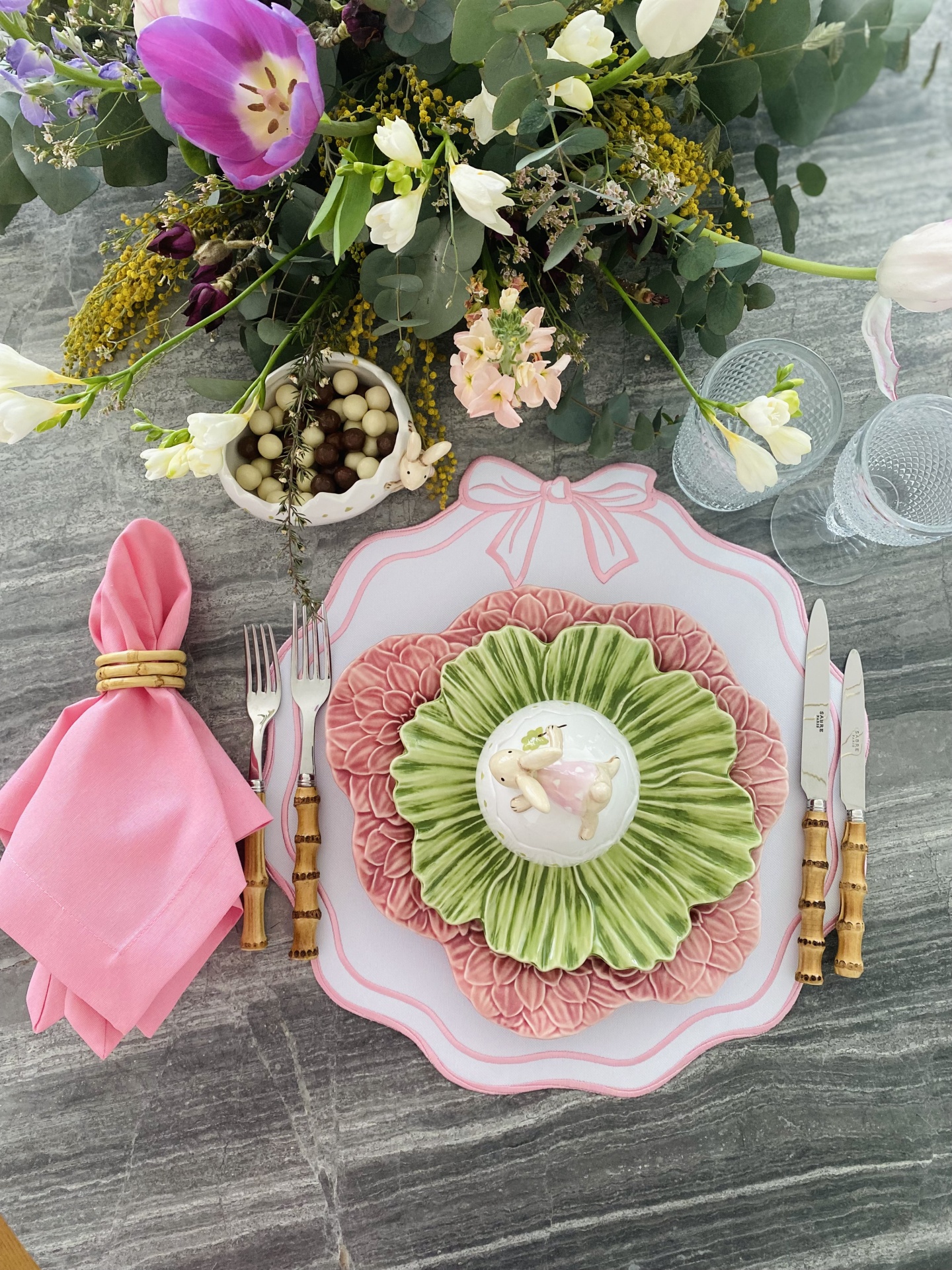 Mesa com pratos decorativos, talheres com cabo imitação bambu, guardanapo rosa, copos e flores