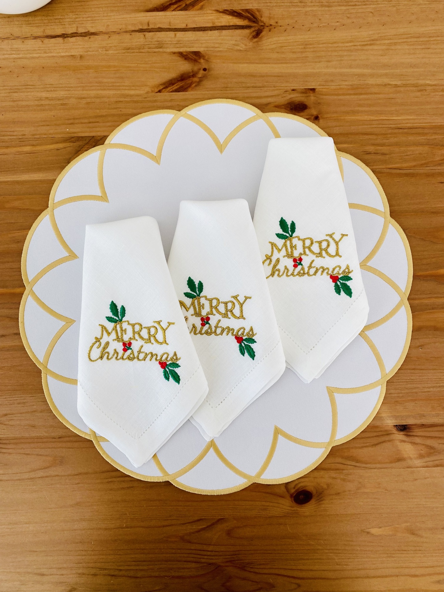 Três guardanapos brancos bordados com texto de Natal sobre prato branco com detalhes dourados
