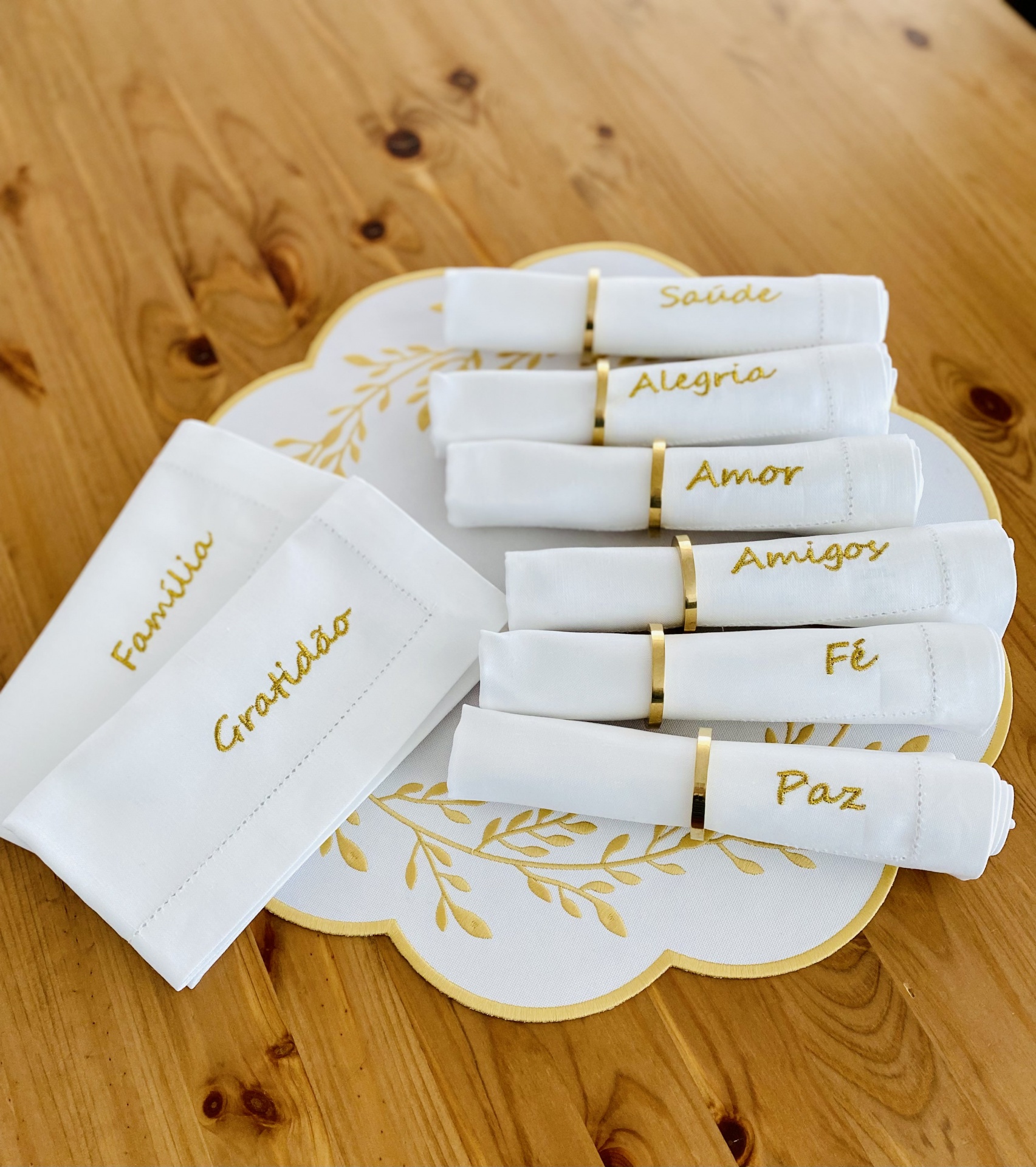 Conjunto de guardanapos brancos com palavras bordadas em dourado e suporte decorativo sobre mesa de madeira