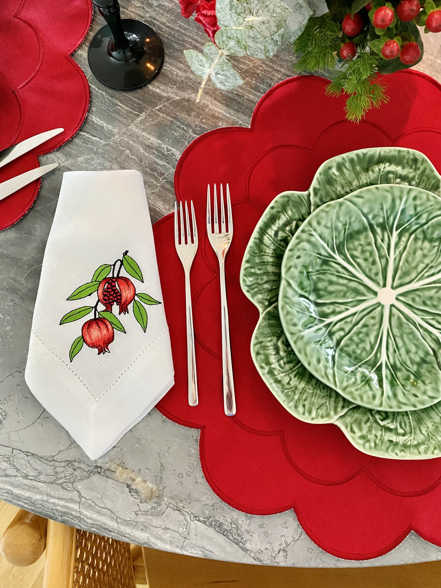 Mesa posta com pratos verdes e vermelhos, guardanapo branco bordado com romãs e talheres de metal