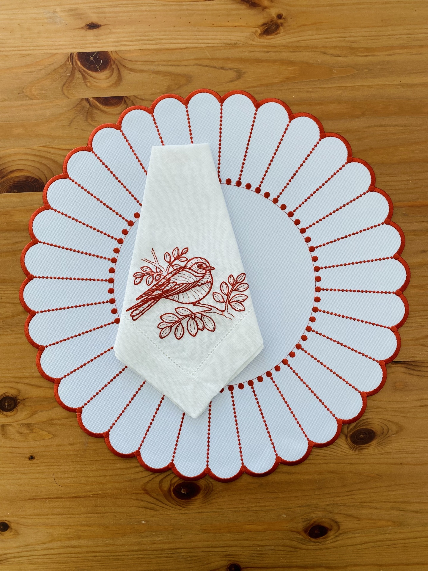 Individual redondo branco com borda vermelha e guardanapo branco bordado com pássaro vermelho sobre mesa de madeira