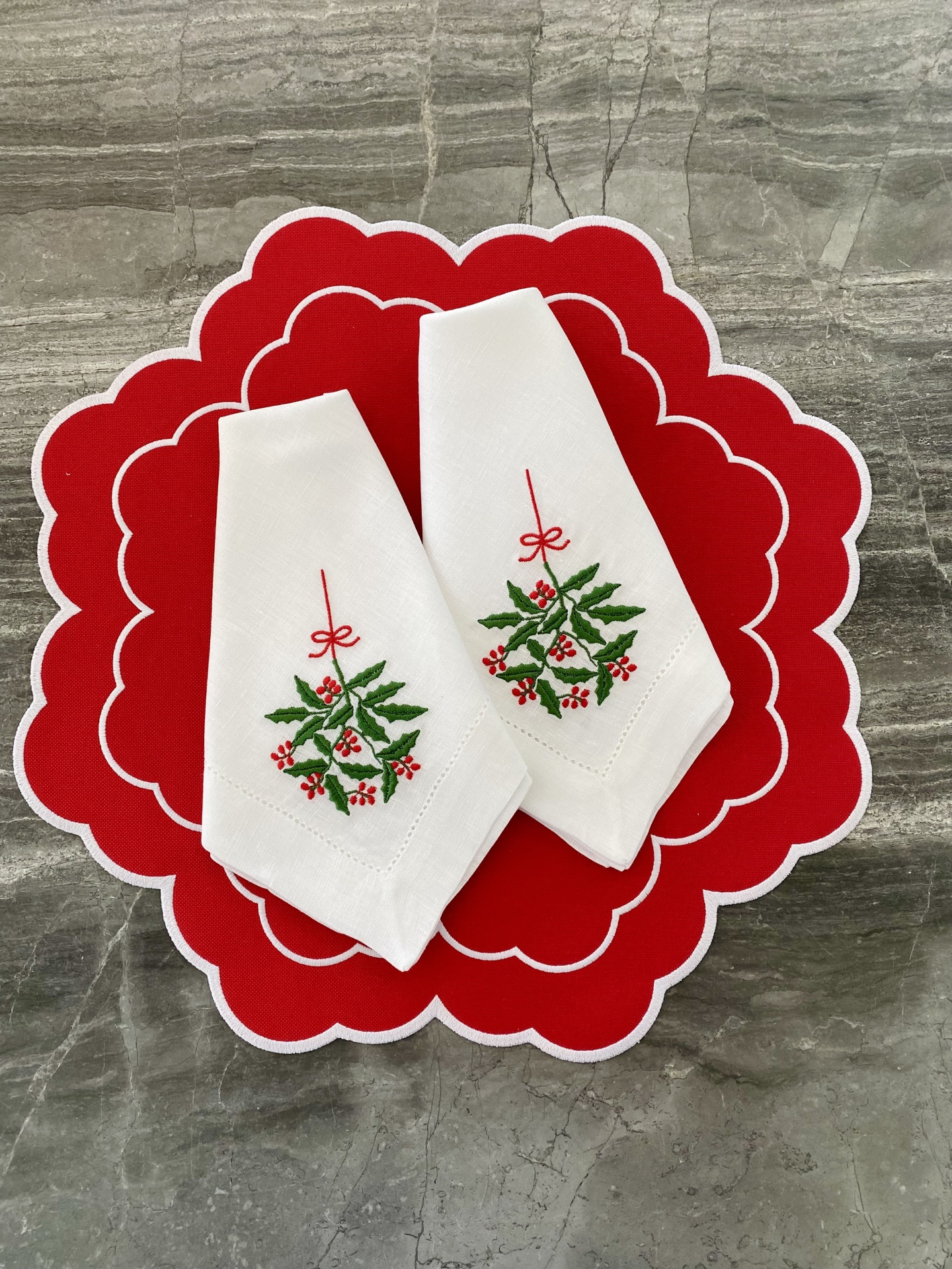 Dois guardanapos brancos bordados com azevinho sobre sousplat vermelho com pétalas brancas numa superfície cinzenta.