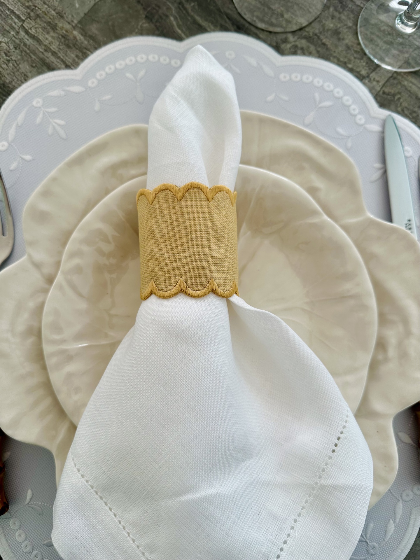 Conjunto de pratos de cerâmica bege com guardanapo branco e argola amarela em mesa de madeira.