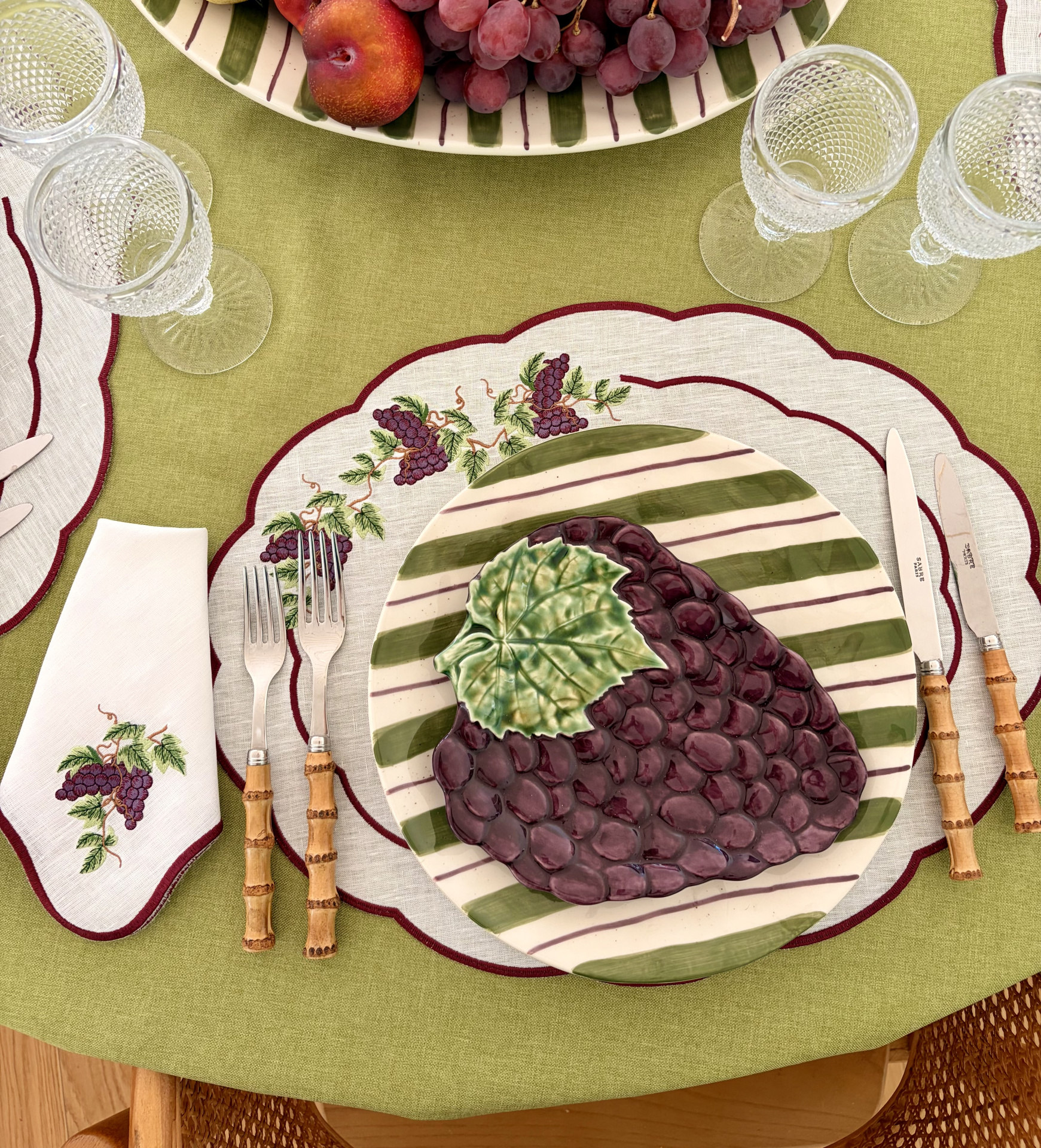 Mesa com toalha verde, prato de cerâmica decorado com cachos de uvas, talheres com cabo de madeira e copos de vidro texturizados