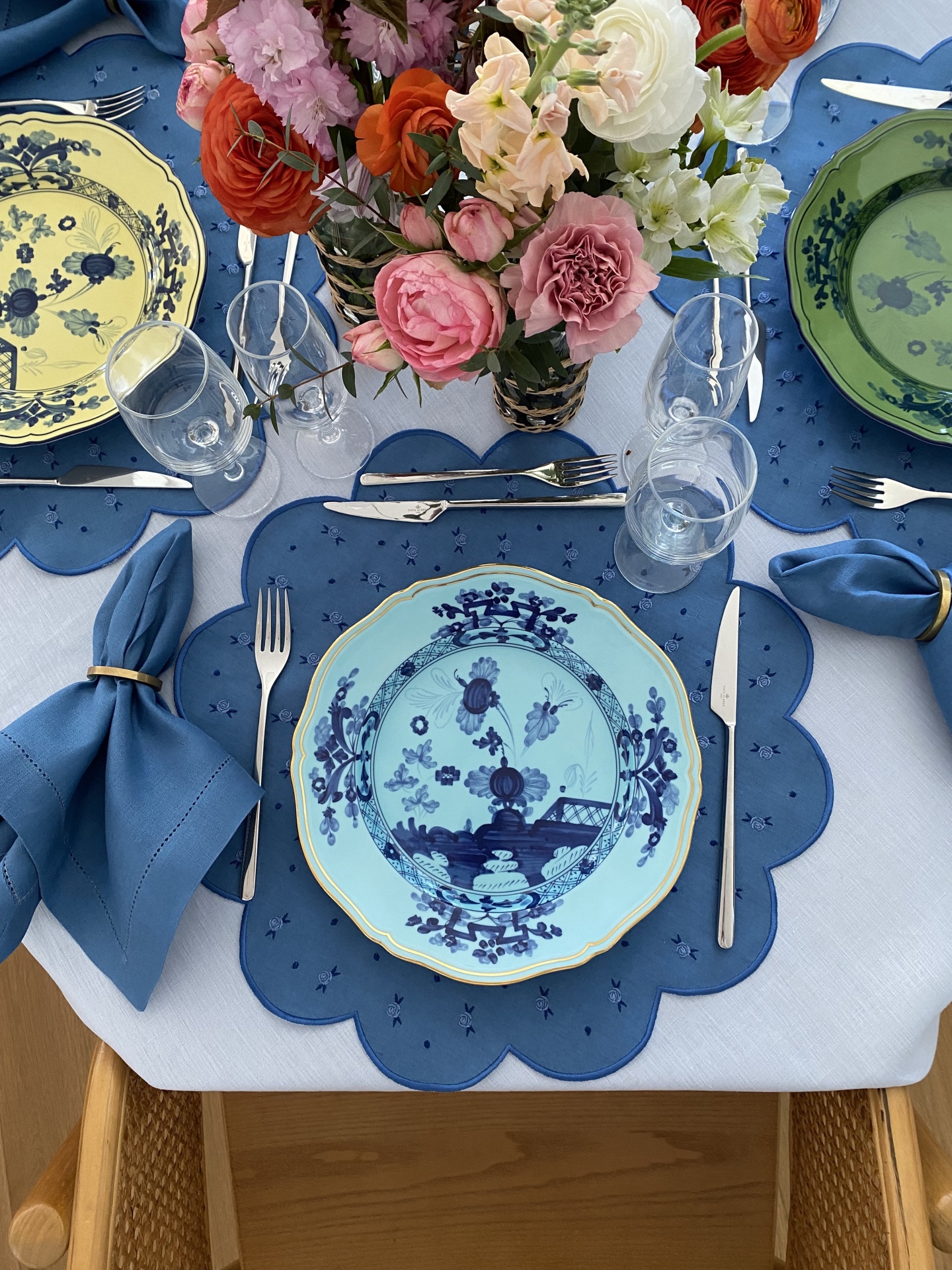 Mesa posta com prato azul claro e azul escuro, individual azul, guardanapos azuis com argolas douradas, talheres e copos de vidro, flores coloridas no centro