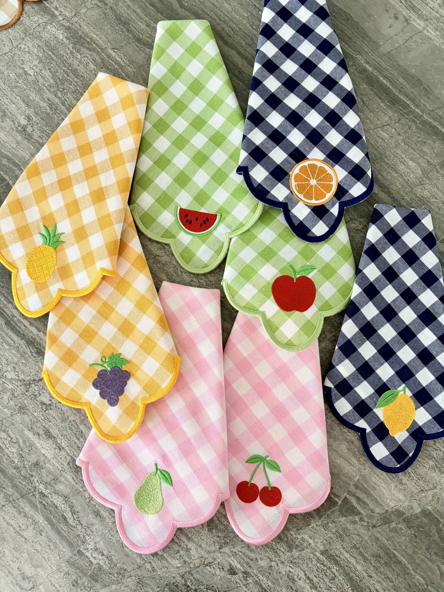 Toalhas de tecido xadrez com bordados de frutas alinhadas numa superfície de pedra cinzenta.
