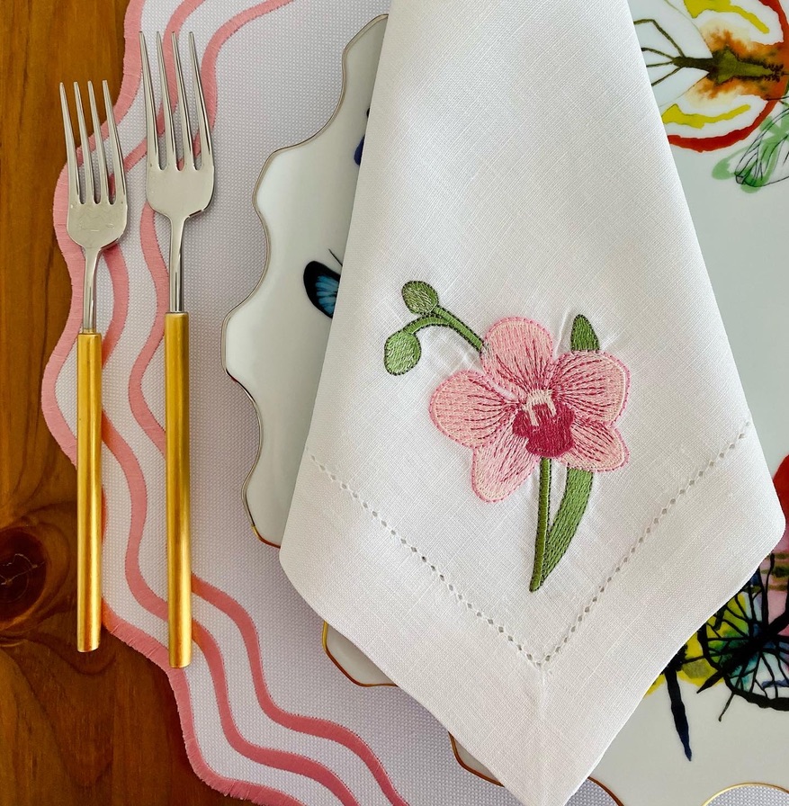 Guardanapo branco com bordado rosa sobre prato com borboletas, jogo americano rosa, garfos com cabo amarelo