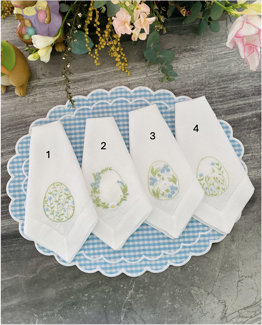 Quatro guardanapos brancos bordados com flores, numerados 1 a 4, sobre tabuleiro azul e branco xadrez em superfície cinzenta