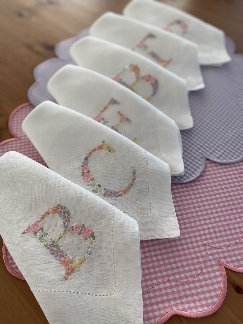 Seis guardanapos brancos bordados com letras florais sobre pano de mesa rosa xadrez e pano lilás
