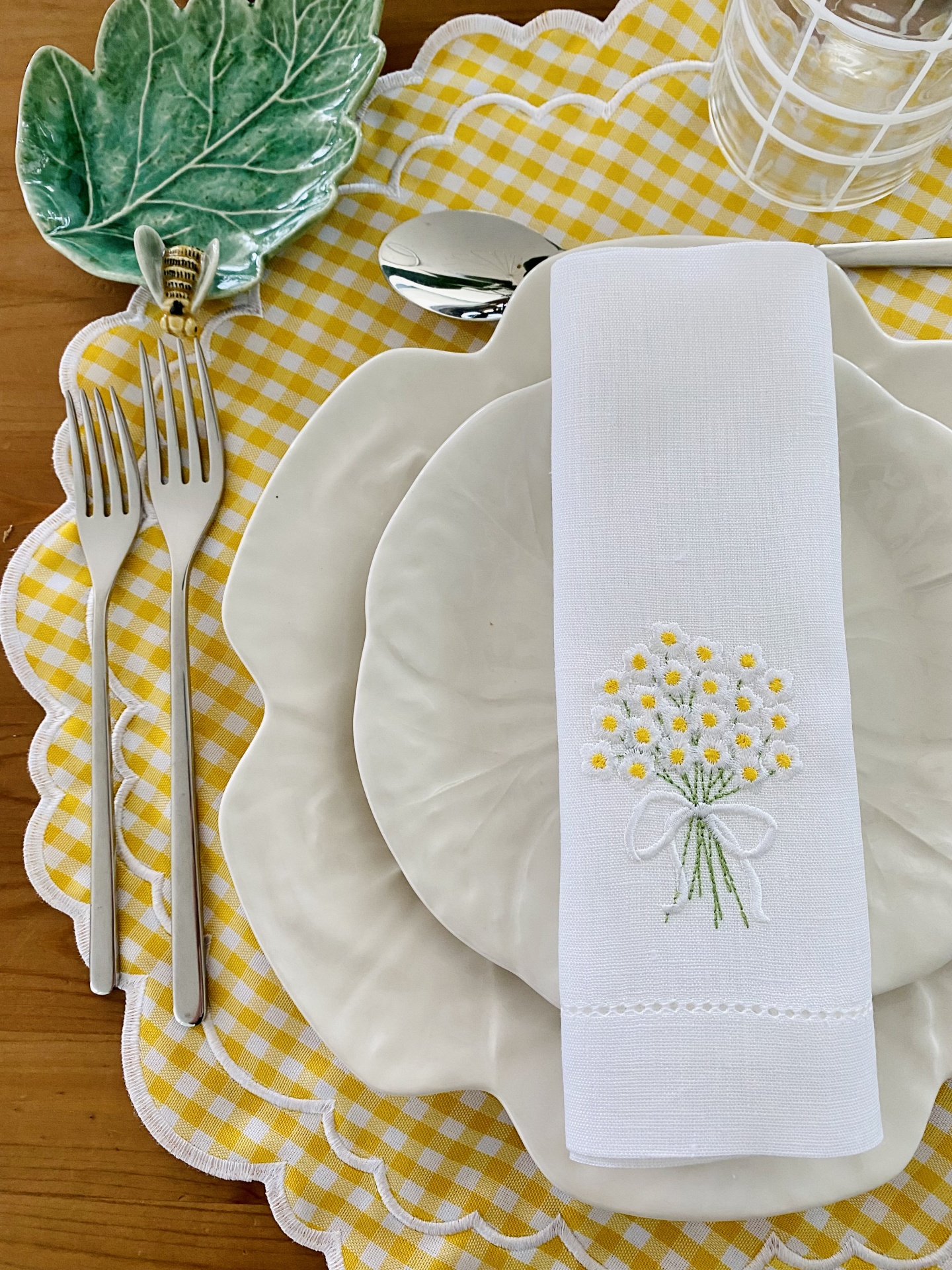 Conjunto de jantar com pratos brancos ondulados, guardanapo bordado, talheres prateados, prato verde em forma de folha e toalha de mesa xadrez amarela