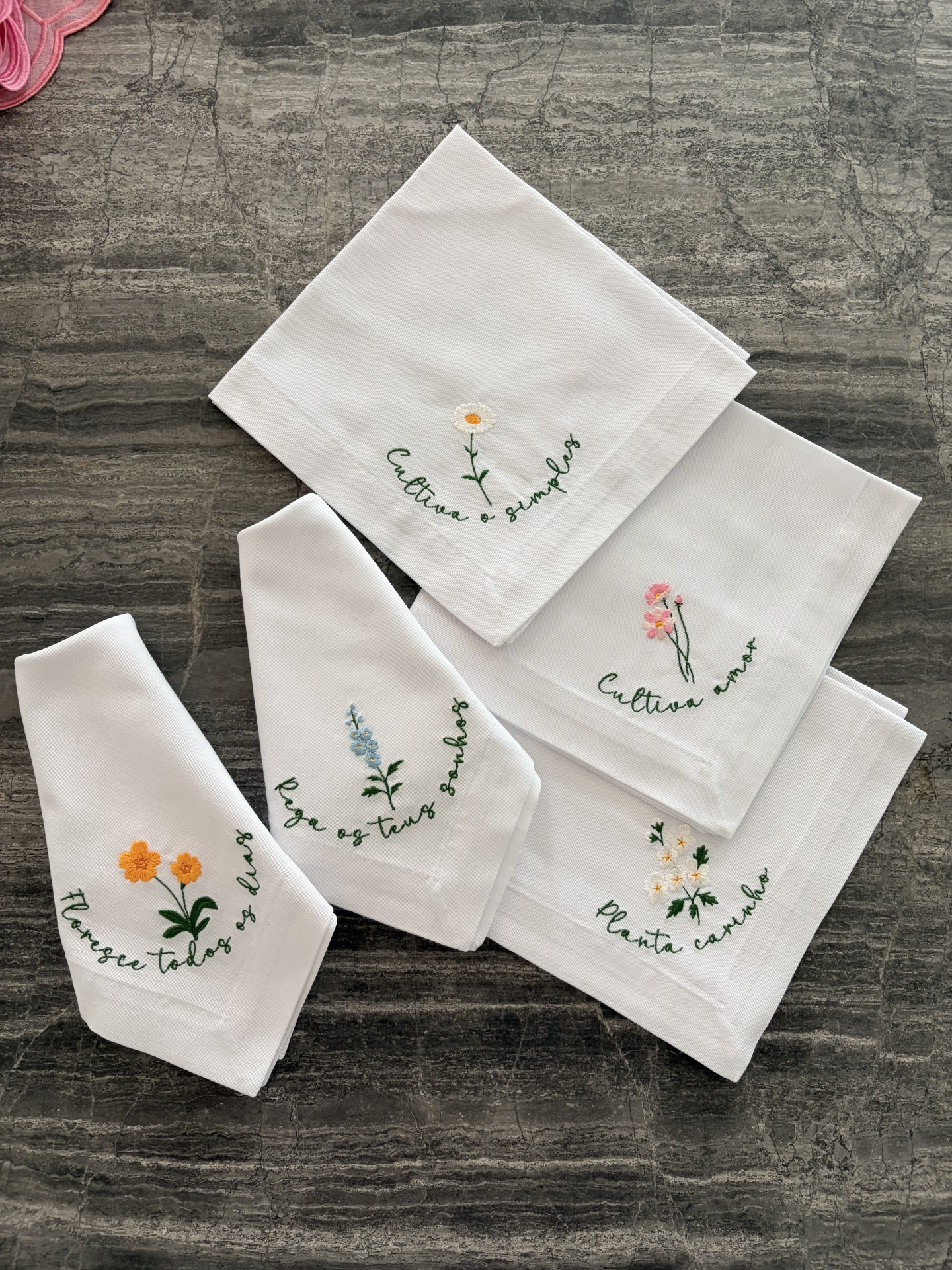 Guardanapos brancos com bordados de flores coloridas e frases em verde sobre superfície cinza.