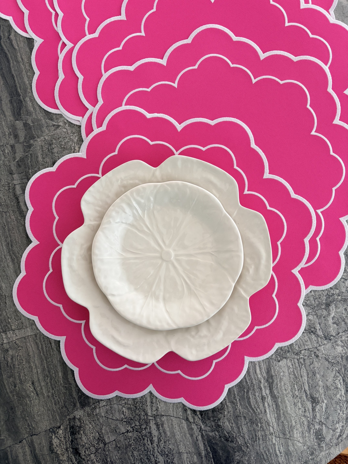 Três pratos brancos em forma de flor sobre descanso de pratos rosa em superfície de pedra.