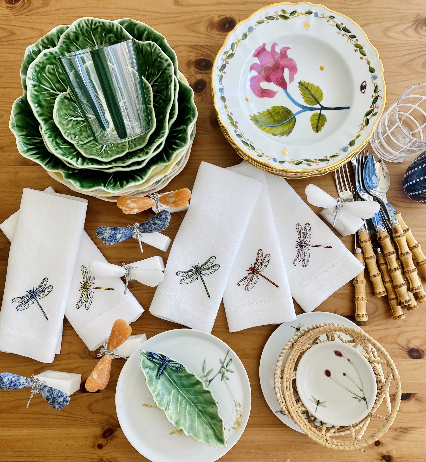 Mesa com pratos cerâmicos decorativos, guardanapos brancos com libélulas bordadas e talheres com cabos castanhos.