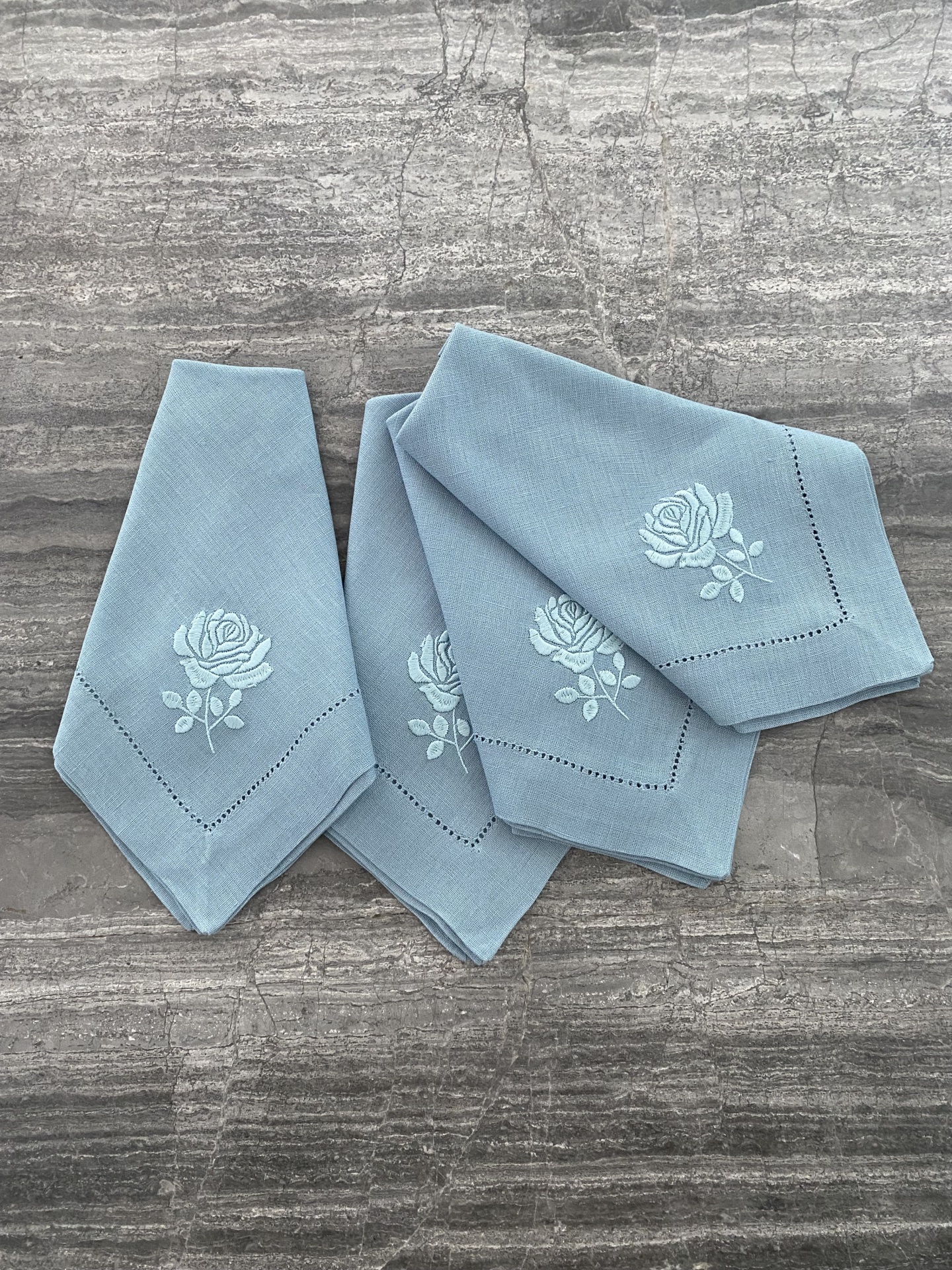 Quatro guardanapos azuis claros com rosa bordada em branco sobre superfície cinza
