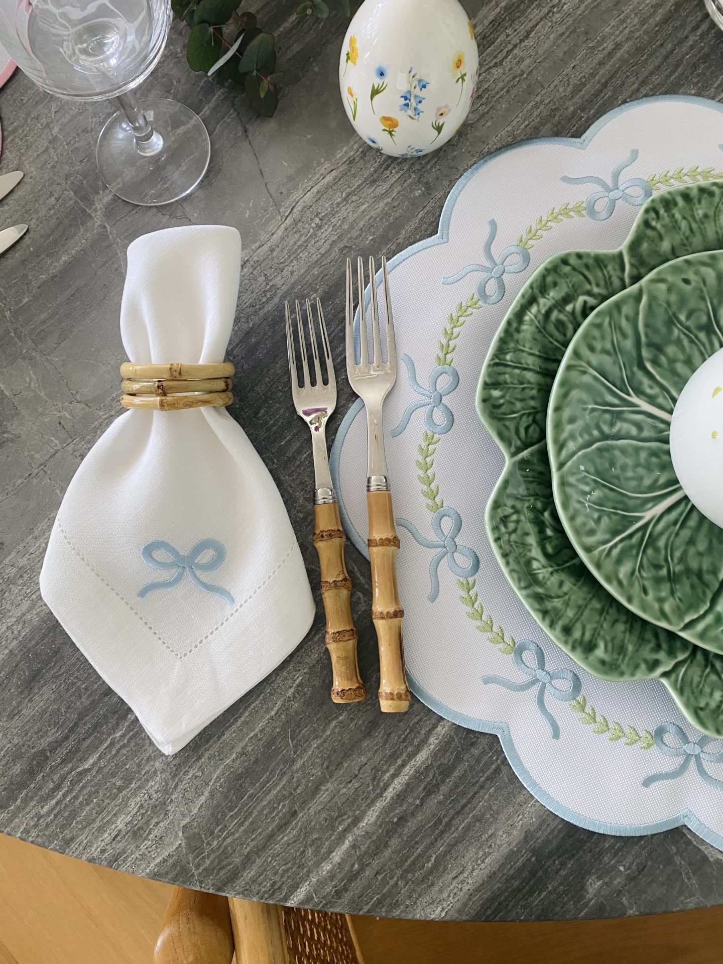 Mesa posta com guardanapo branco, garfos com cabo de bambu, prato verde em forma de folha, base branca com pattern azul, ovo decorativo e copo de vinho