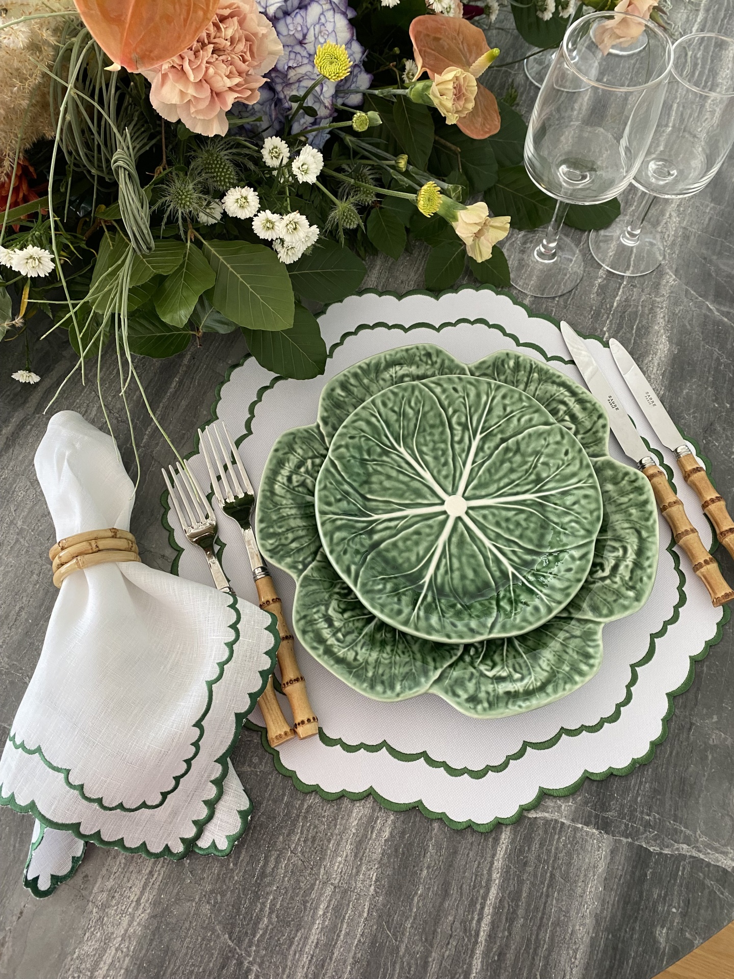 Lugar de mesa com louça verde com relevo de folhas, guardanapo branco, talheres com cabo castanho e copos de vidro numa mesa cinzenta com flores.