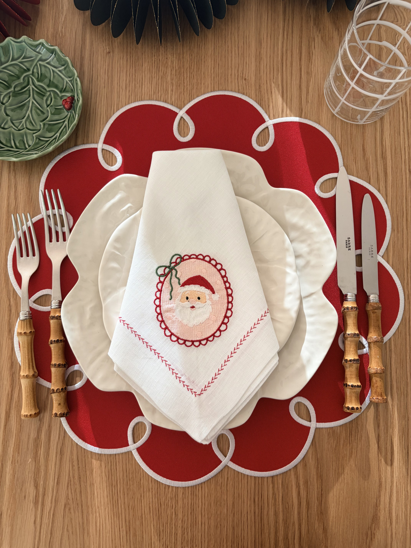 Mesa posta com pratos brancos, guardanapo com bordado de Pai Natal, talheres com cabos de bambu e placemat vermelho