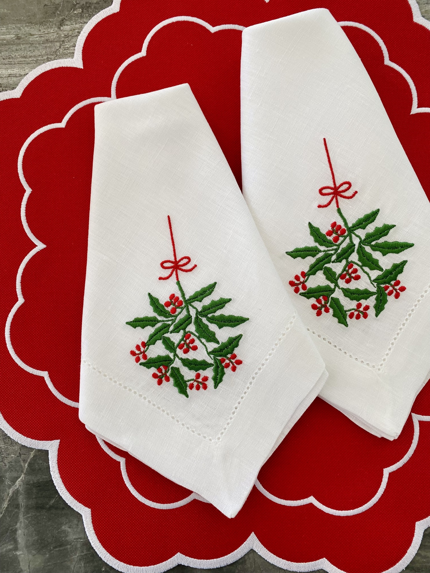 Duas guardanapos brancos bordados com azevinho sobre base vermelha com contorno branco