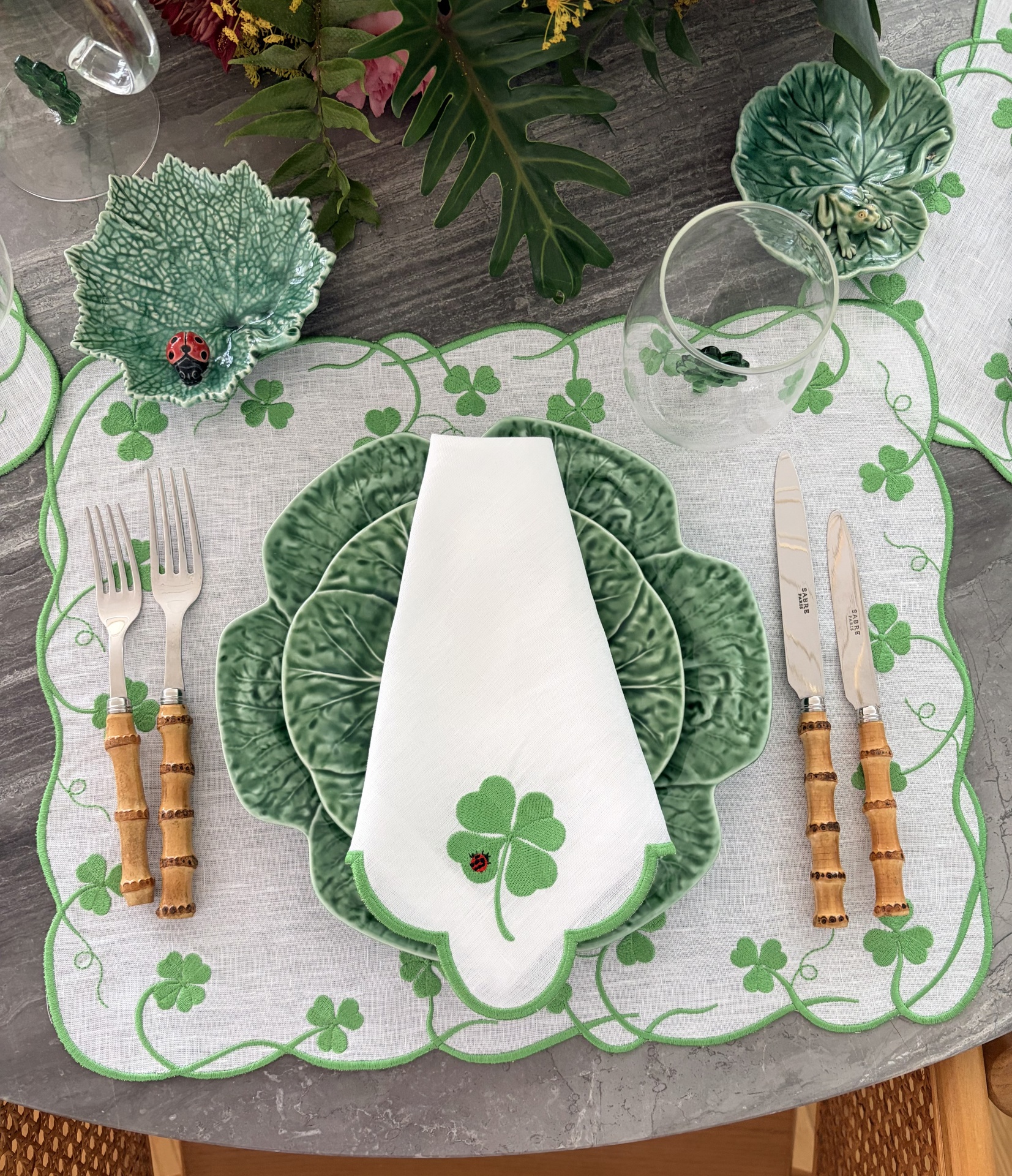 Mesa posta com loiça verde formato de folhas, guardanapo branco com bordado de trevo e joaninha, talheres com cabo de madeira e copo transparente sobre toalha bordada trevos verdes