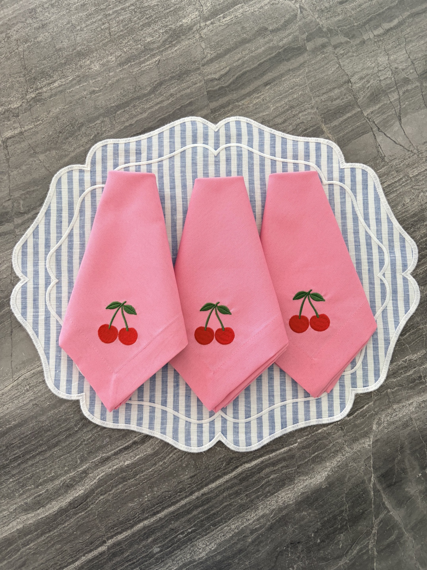Três guardanapos cor-de-rosa com bordado de cerejas sobre suportes de tecido riscado azul e branco em superfície de pedra cinzenta.