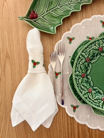Mesa posta com pratos cerâmicos verdes com padrão de folhas, guardanapo branco com argola decorativa, talheres e toalha com estampas natalícias.