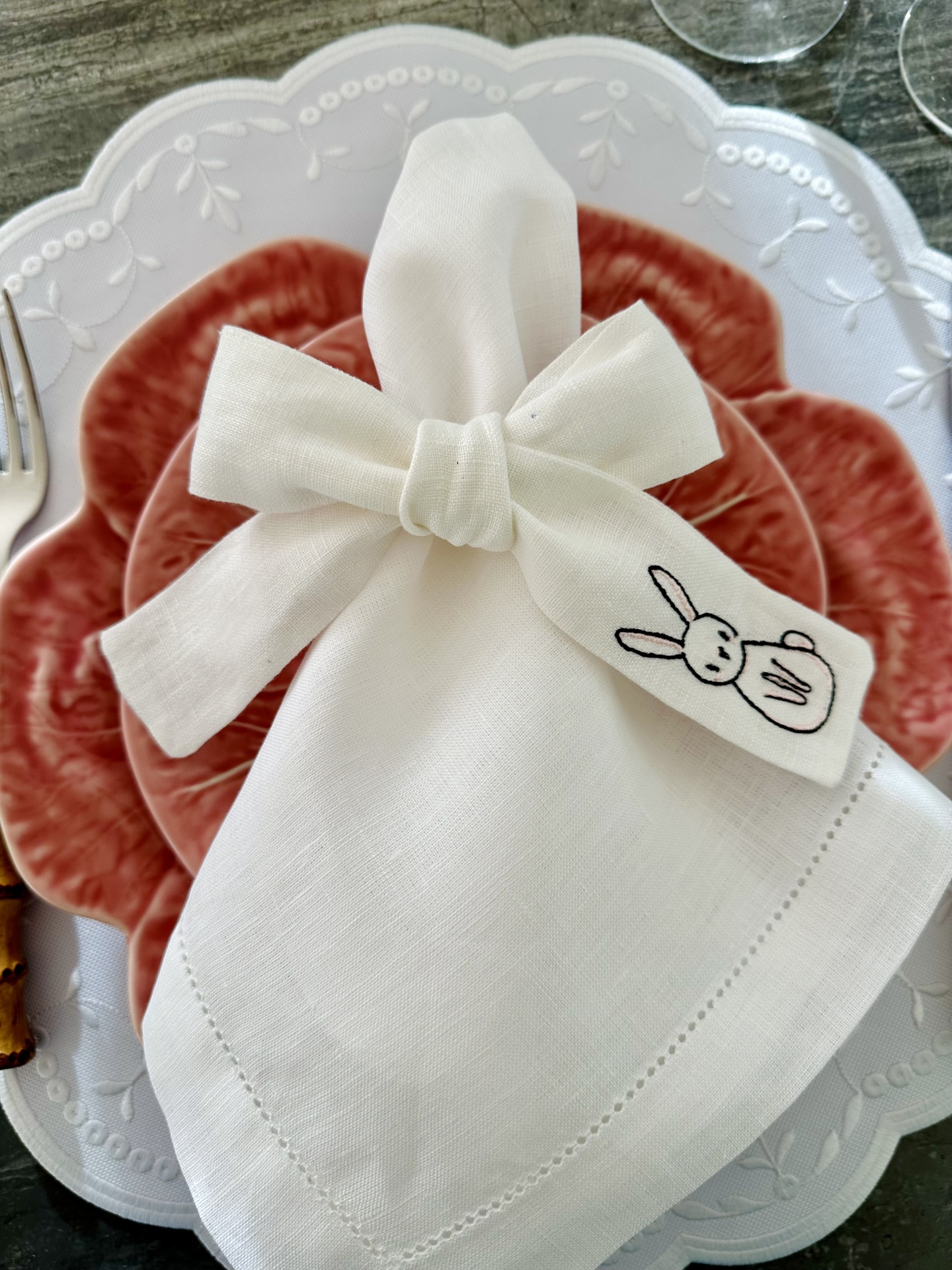 Guardanapo branco com bordado de coelho num prato vermelho e um prato branco por baixo, sobre mesa de madeira