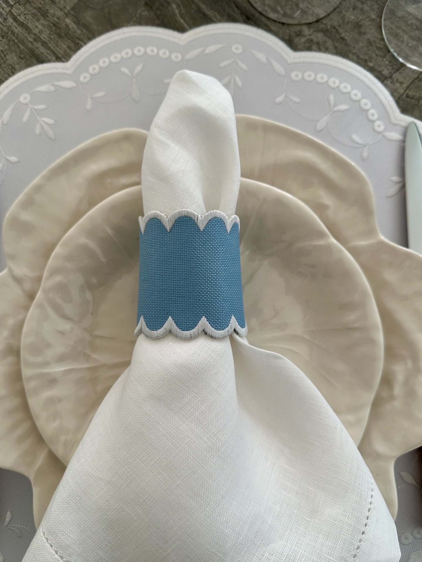 Guardanapo branco com argola azul sobre pratos bege e sousplat branco bordado