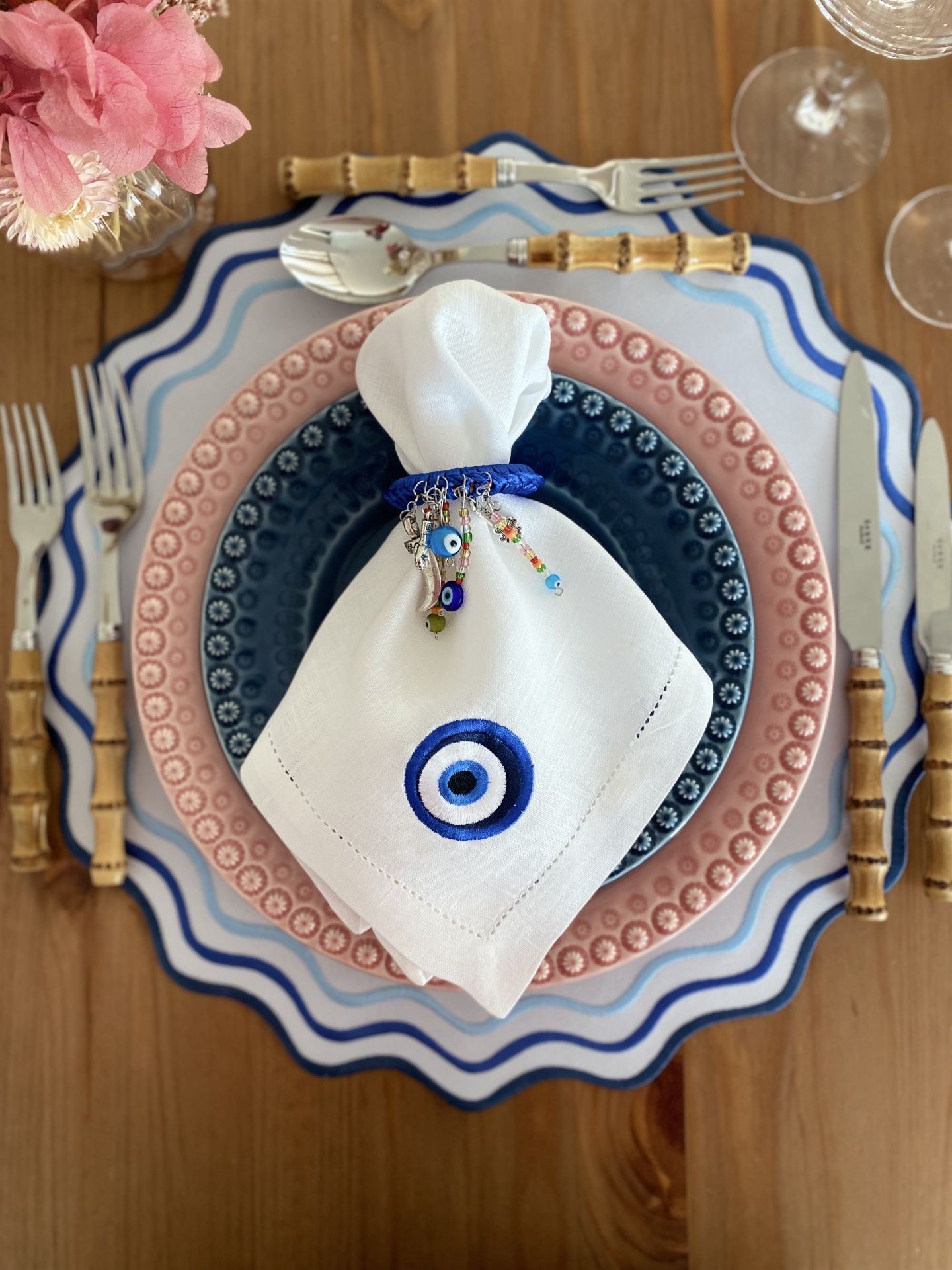 Mesa posta com pratos decorativos azul e rosa, talheres com cabo de bambu, guardanapo branco com olho turco azul e flores cor-de-rosa.