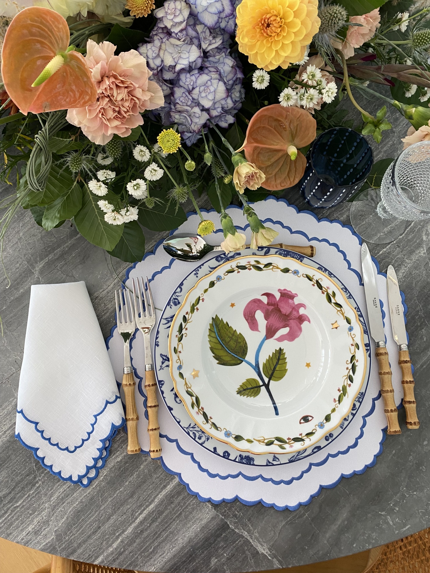 Mesa posta com loiça floral, talheres de cabo de madeira, guardanapo bordado azul e flores mistas