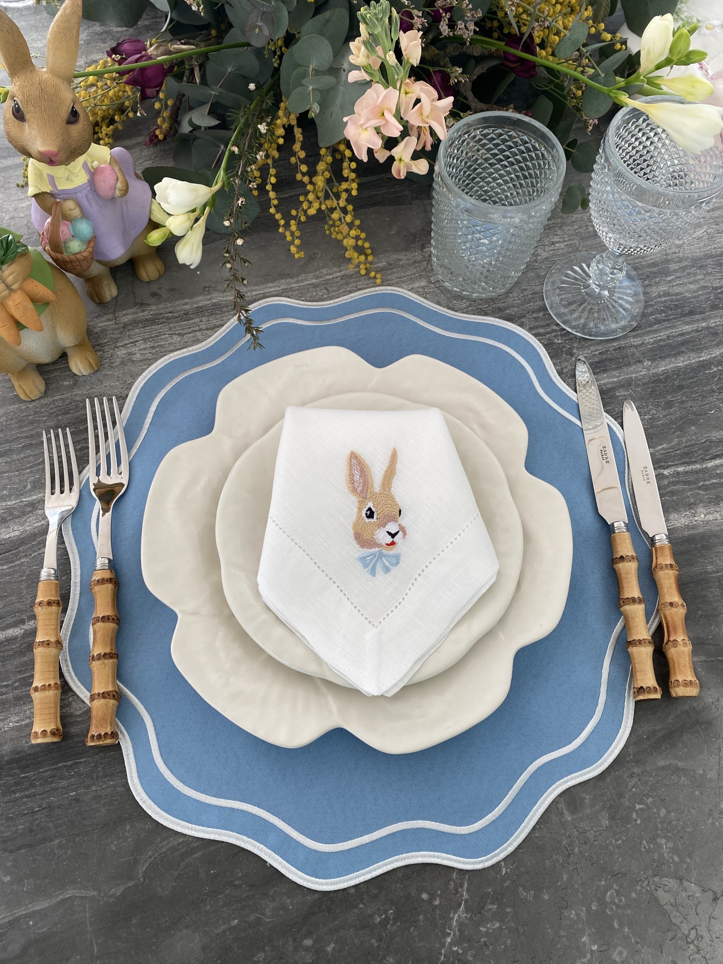 Mesa posta com pratos brancos, guardanapo bordado, talheres com cabo de madeira e decoração de Páscoa